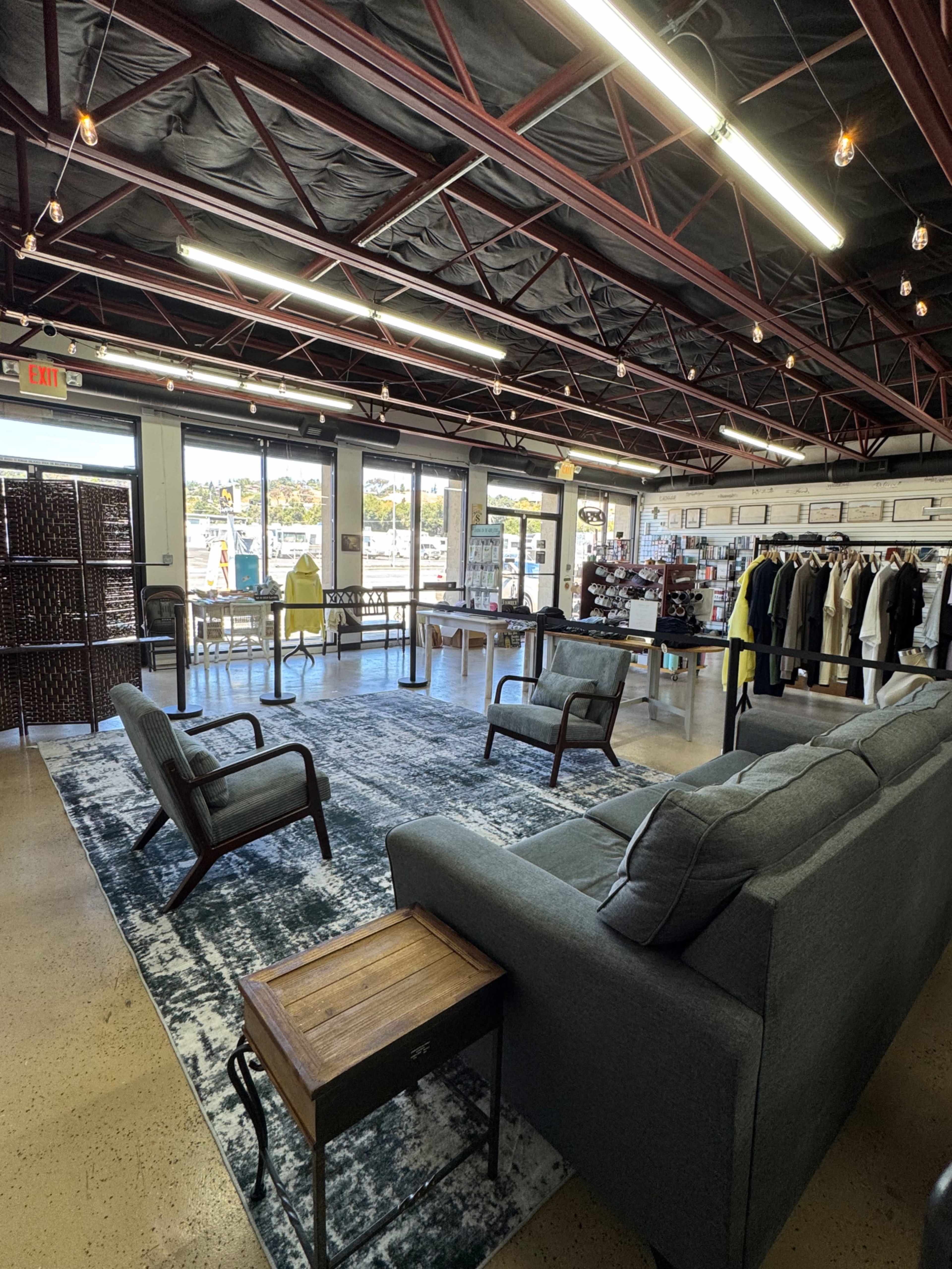 The image shows a modern retail space with a seating area featuring two armchairs and a coffee table on a large area rug, surrounded by clothing racks and large windows letting in natural light.