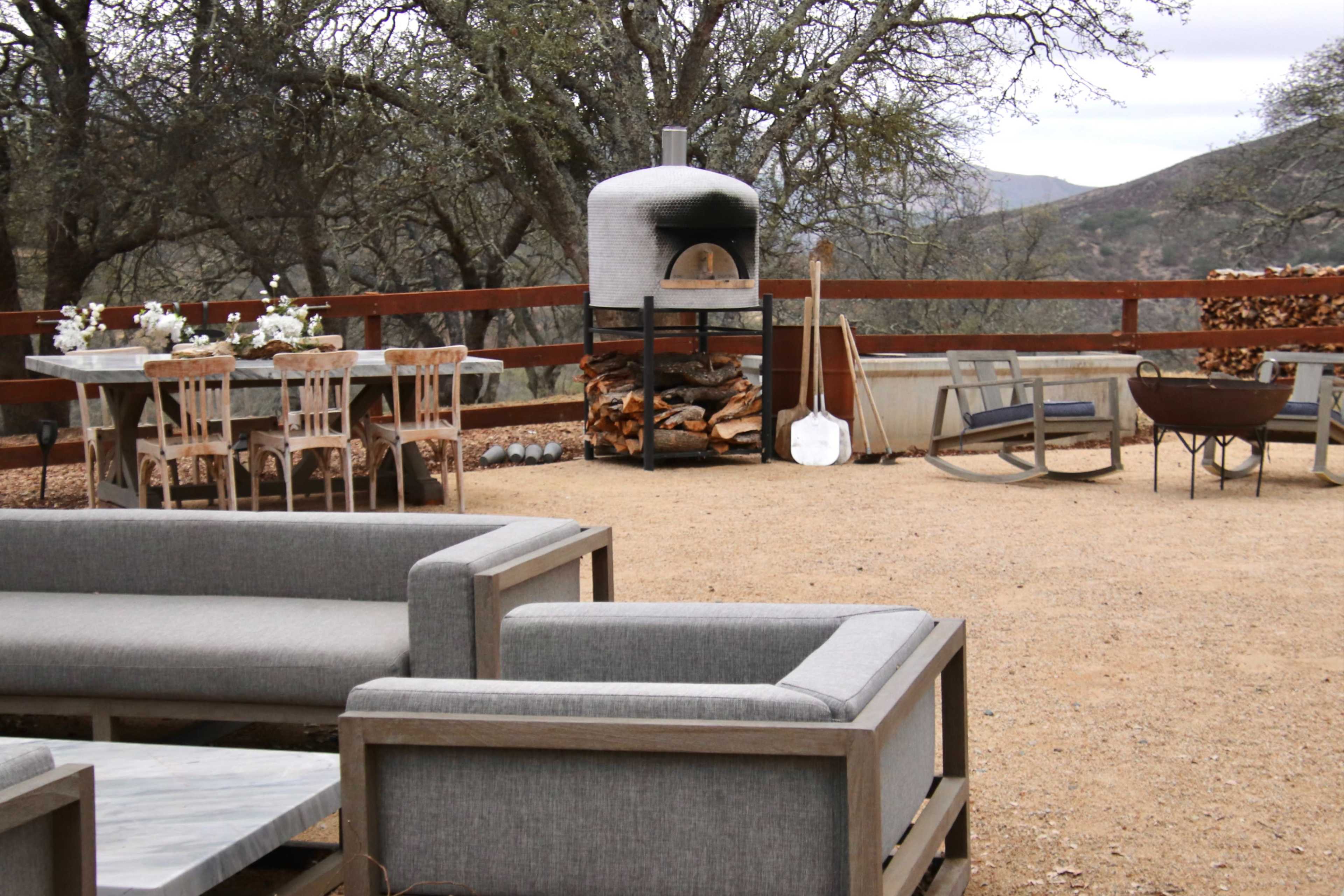 A spacious outdoor patio features a stone pizza oven, a wooden dining table, and several comfortable seating arrangements surrounded by trees and a scenic backdrop.