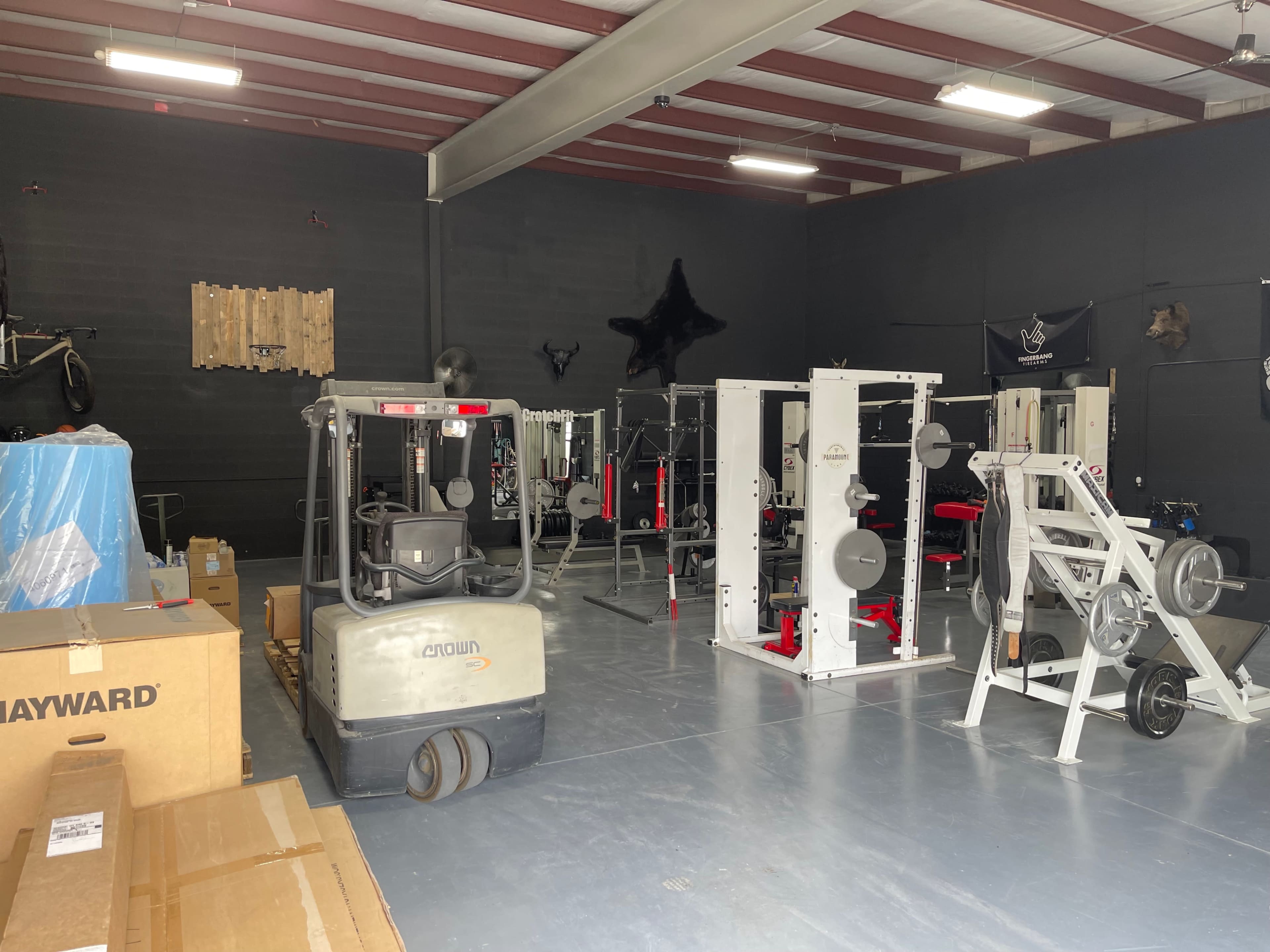 A gym interior with various exercise equipment, boxes, and a forklift in a spacious, industrial-style room.