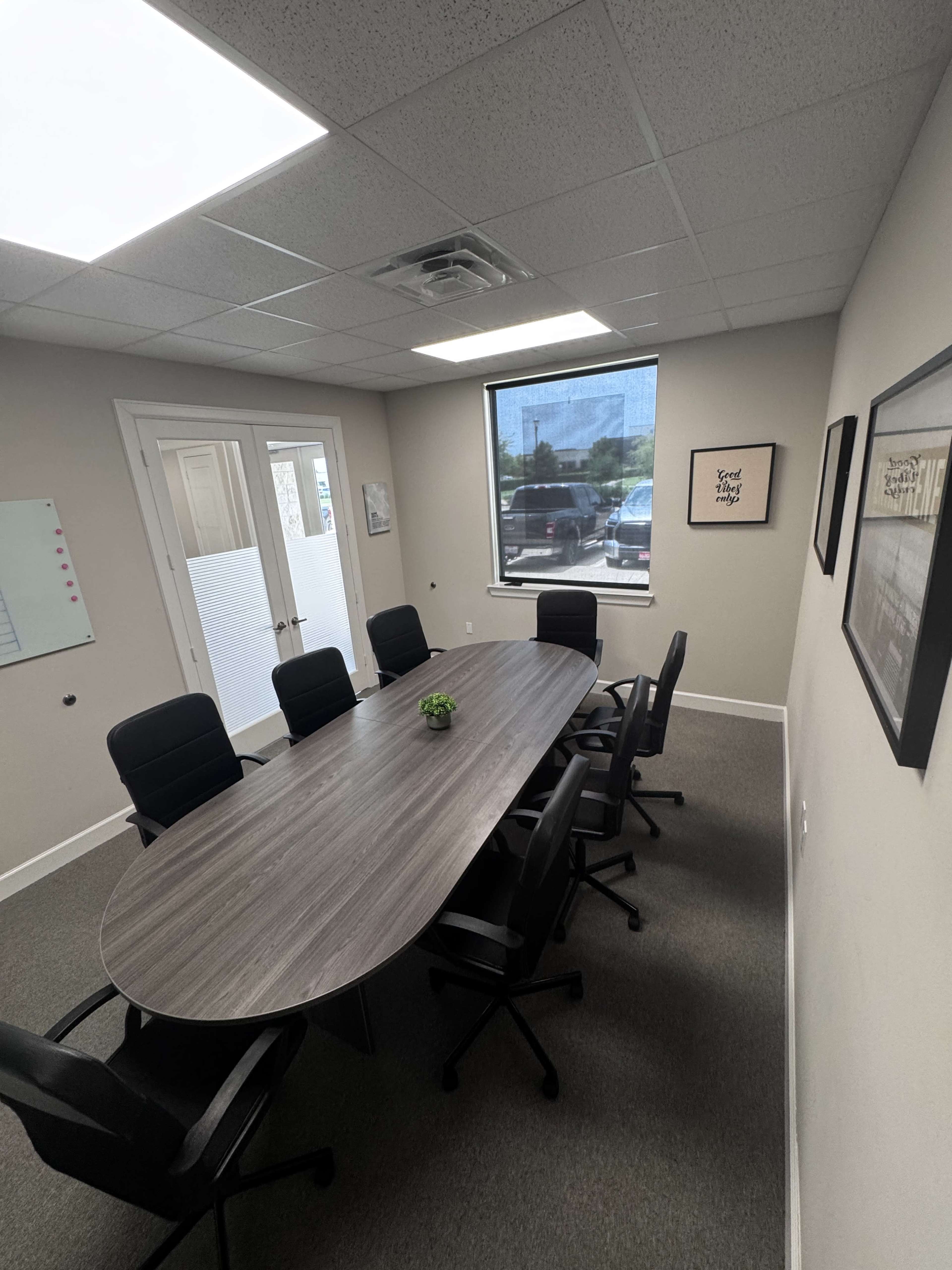 The image shows a conference room with a large oval table surrounded by black chairs, featuring a window with natural light and a whiteboard on the wall.