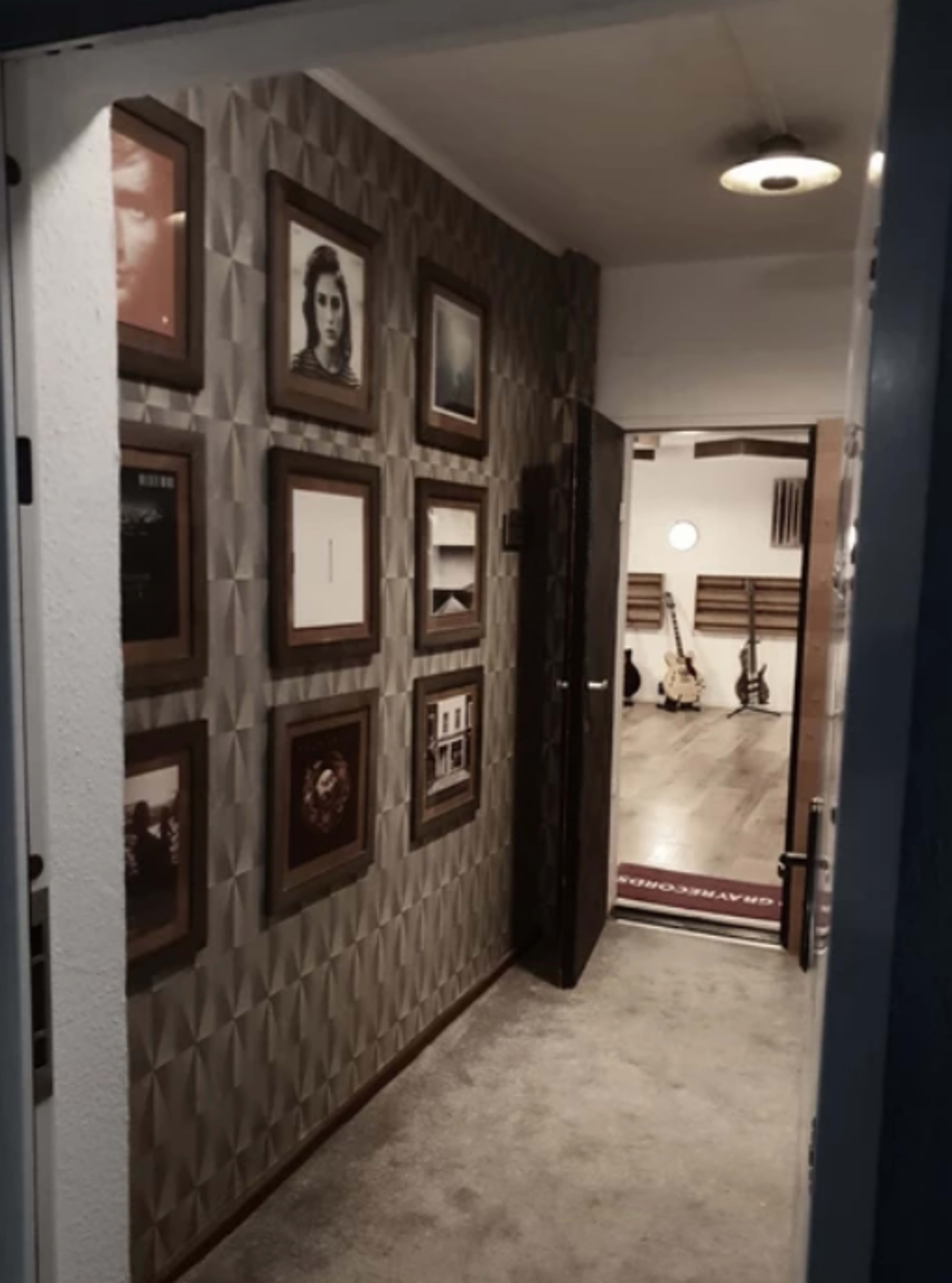 The image shows a hallway with framed pictures on the wall, leading to a spacious room with guitars and a circular light fixture.