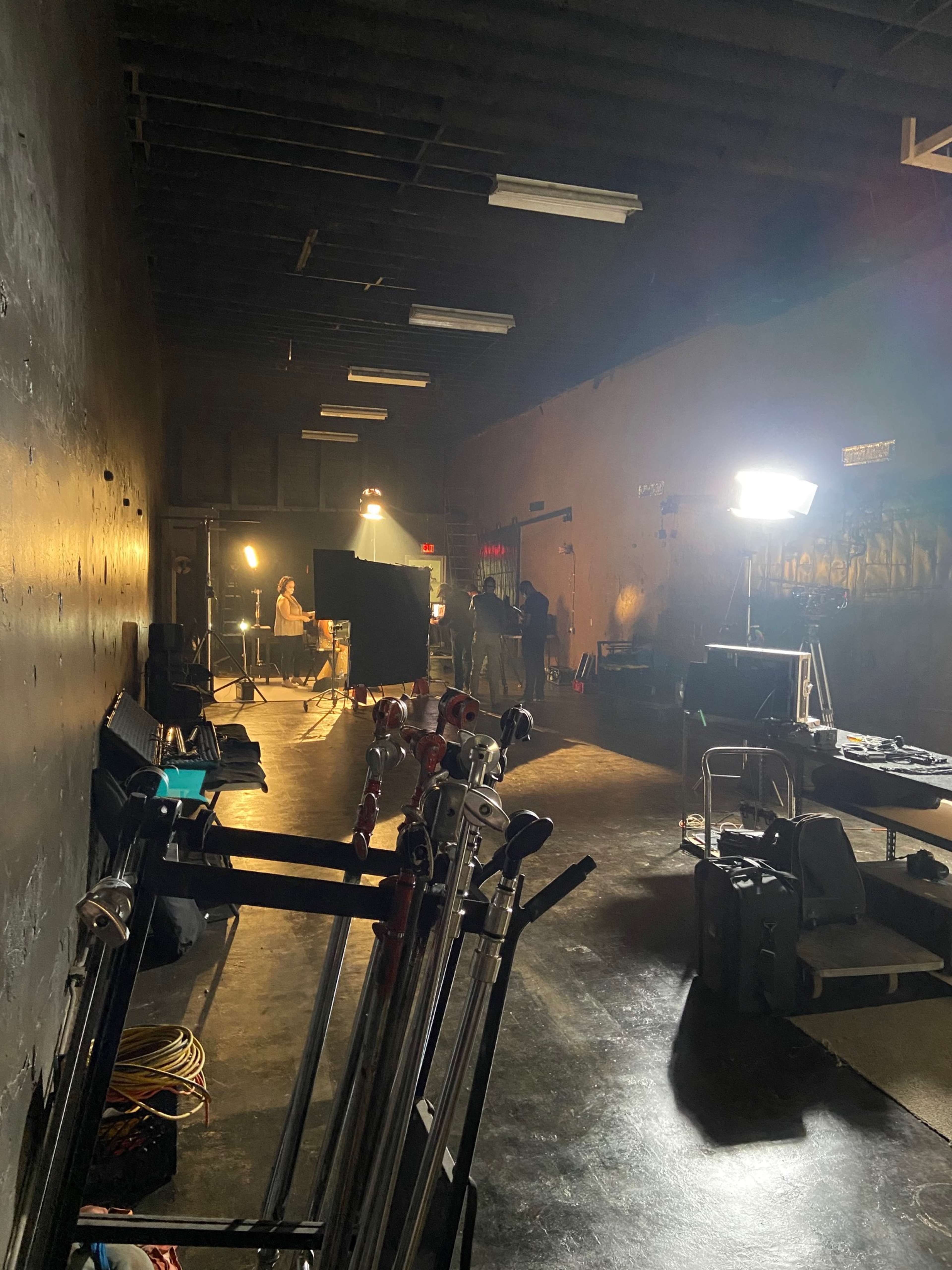 The image shows a dimly lit studio space with several lighting setups and equipment positioned along the left wall.