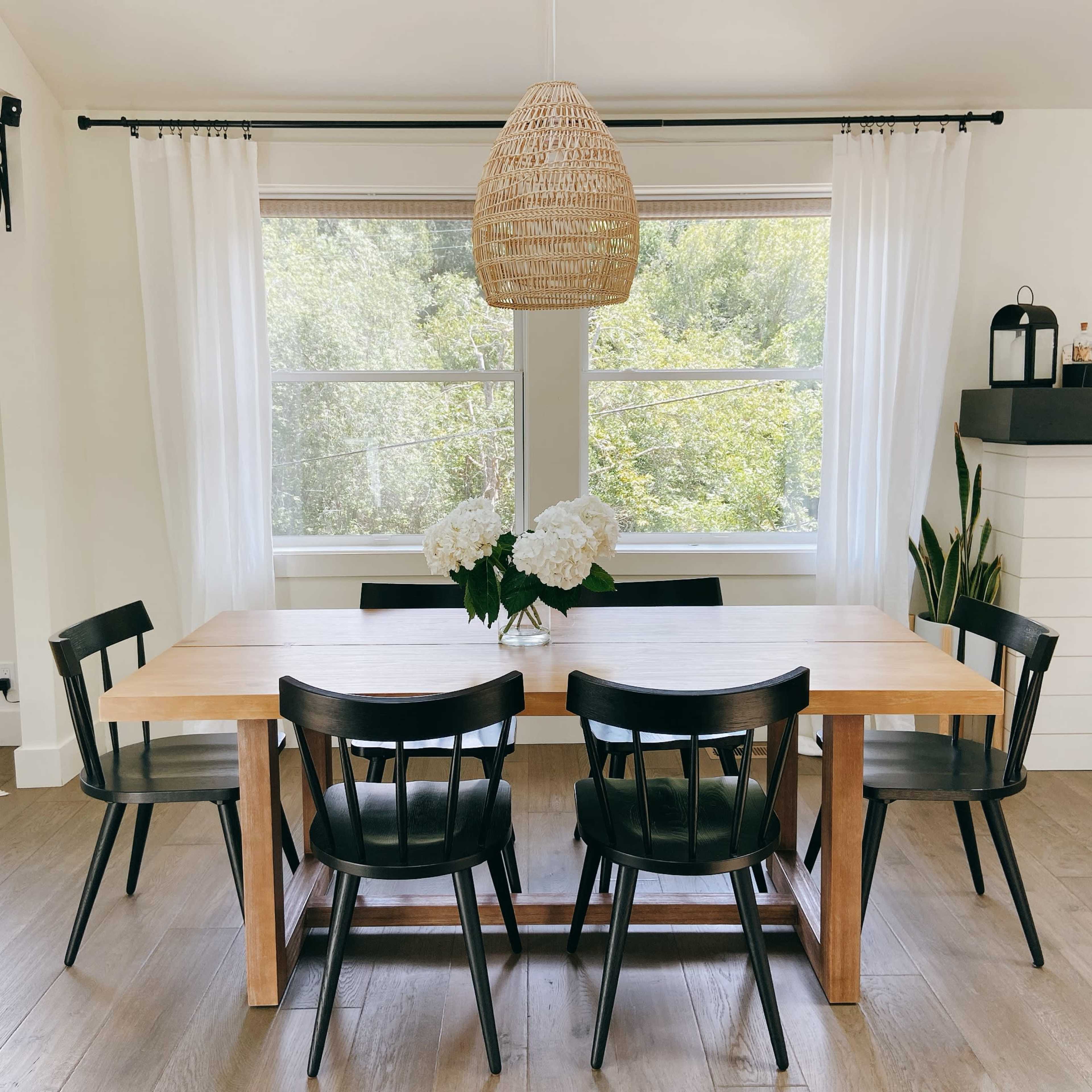 A wooden dining table with black chairs is set in a bright room with large windows and a pendant light overhead, accompanied by a vase of flowers.