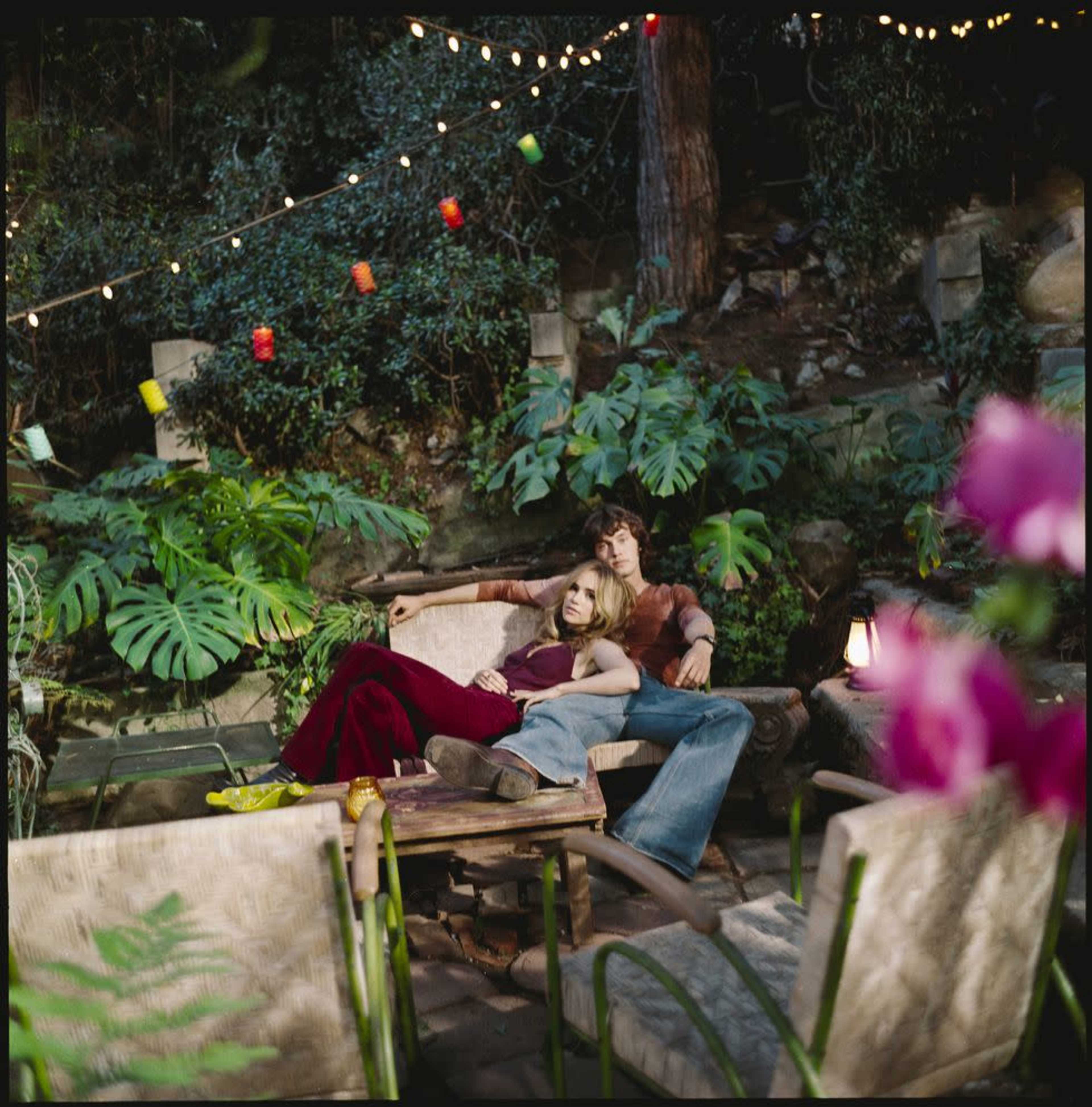 A couple sits closely together on a wooden bench in a lush garden illuminated by string lights.