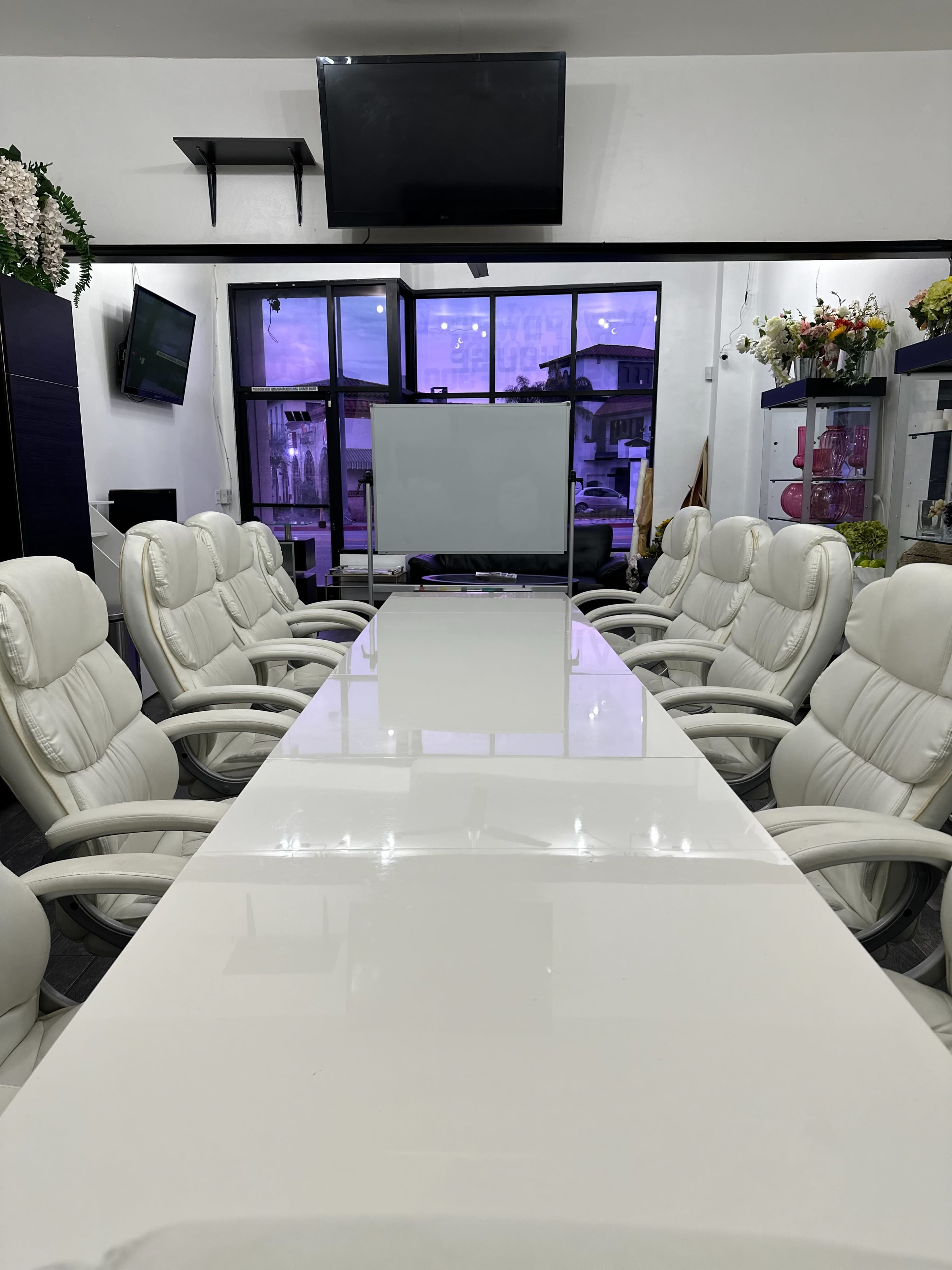 A long, white conference table surrounded by ergonomic chairs is set up in a room with large windows and a wall-mounted television.