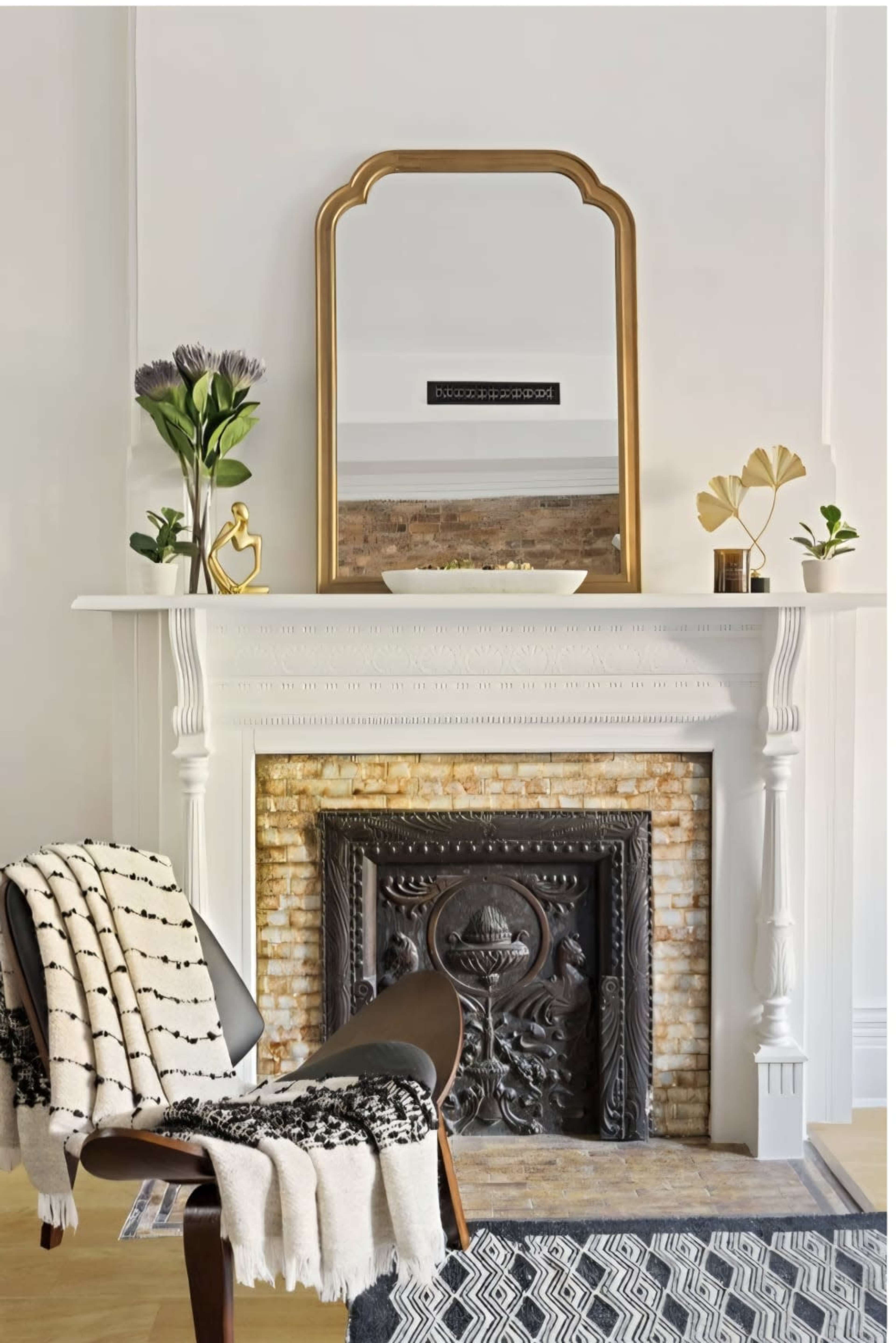 A decorative fireplace with an ornate design sits beneath a gold-framed mirror, surrounded by minimalistic decor including a plant and a small sculpture on the mantel.