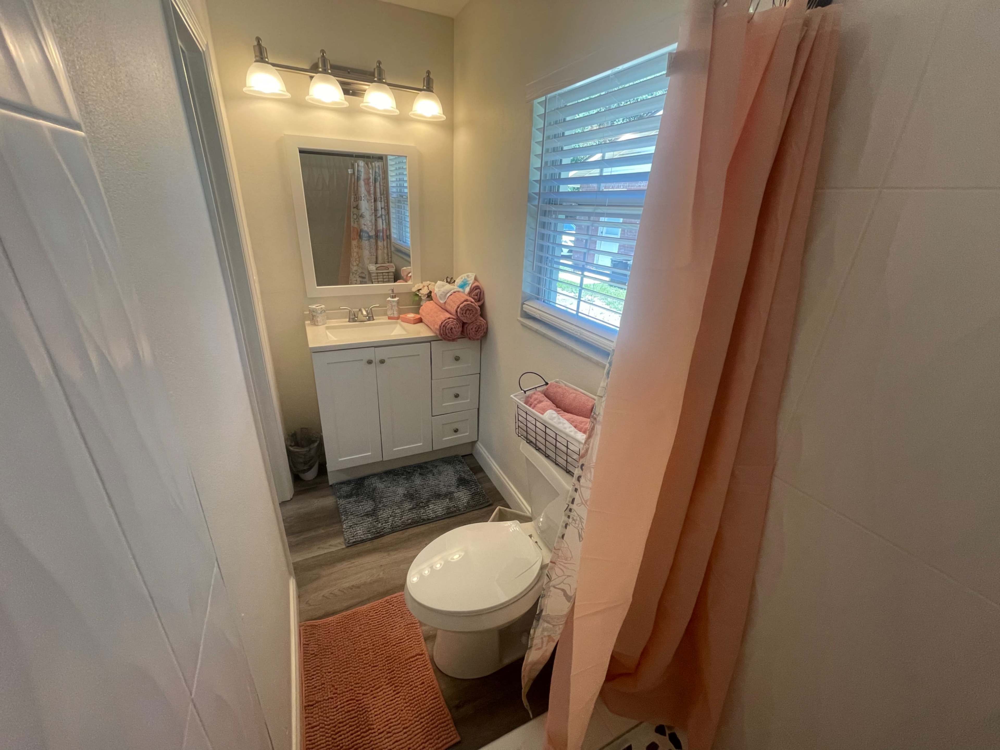 A small bathroom featuring a white vanity with a mirror, a toilet, and a shower curtain, along with neatly arranged towels and a window.
