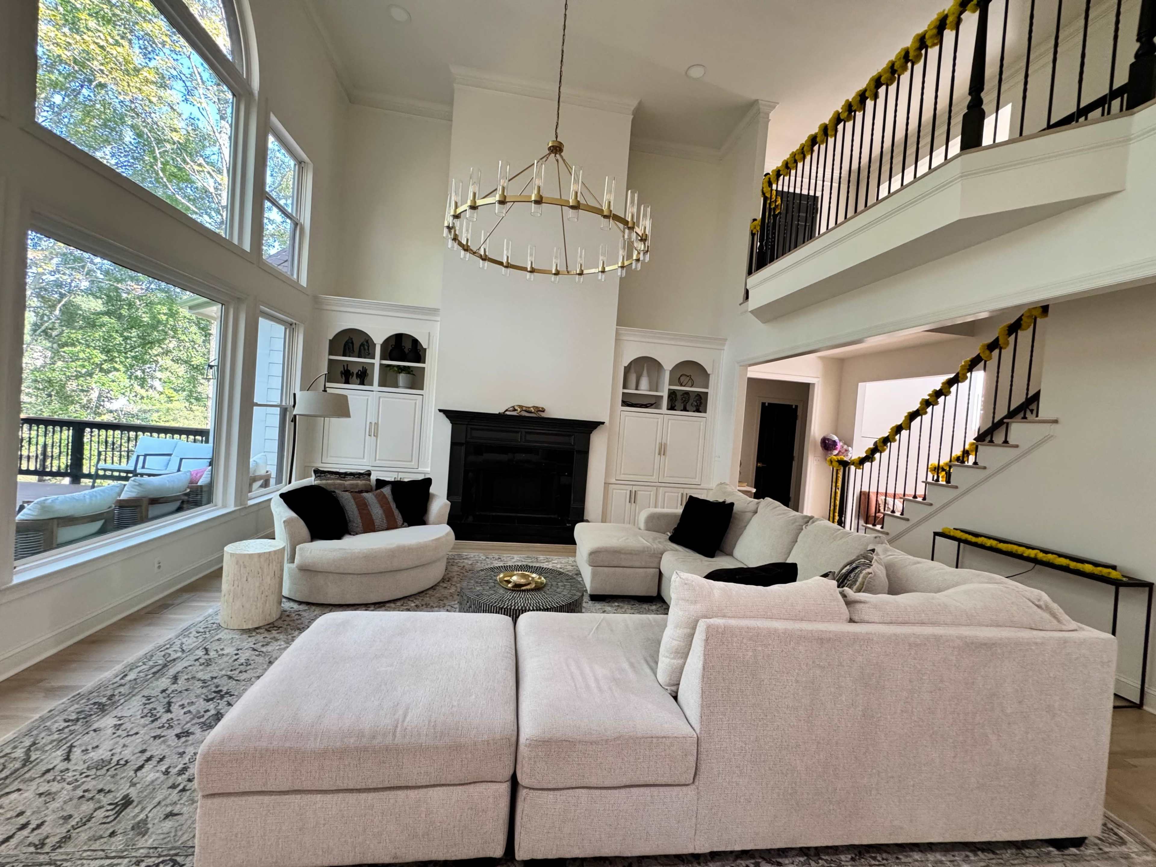 The image depicts a spacious living room with high ceilings, large windows, and a central seating arrangement surrounding a coffee table.