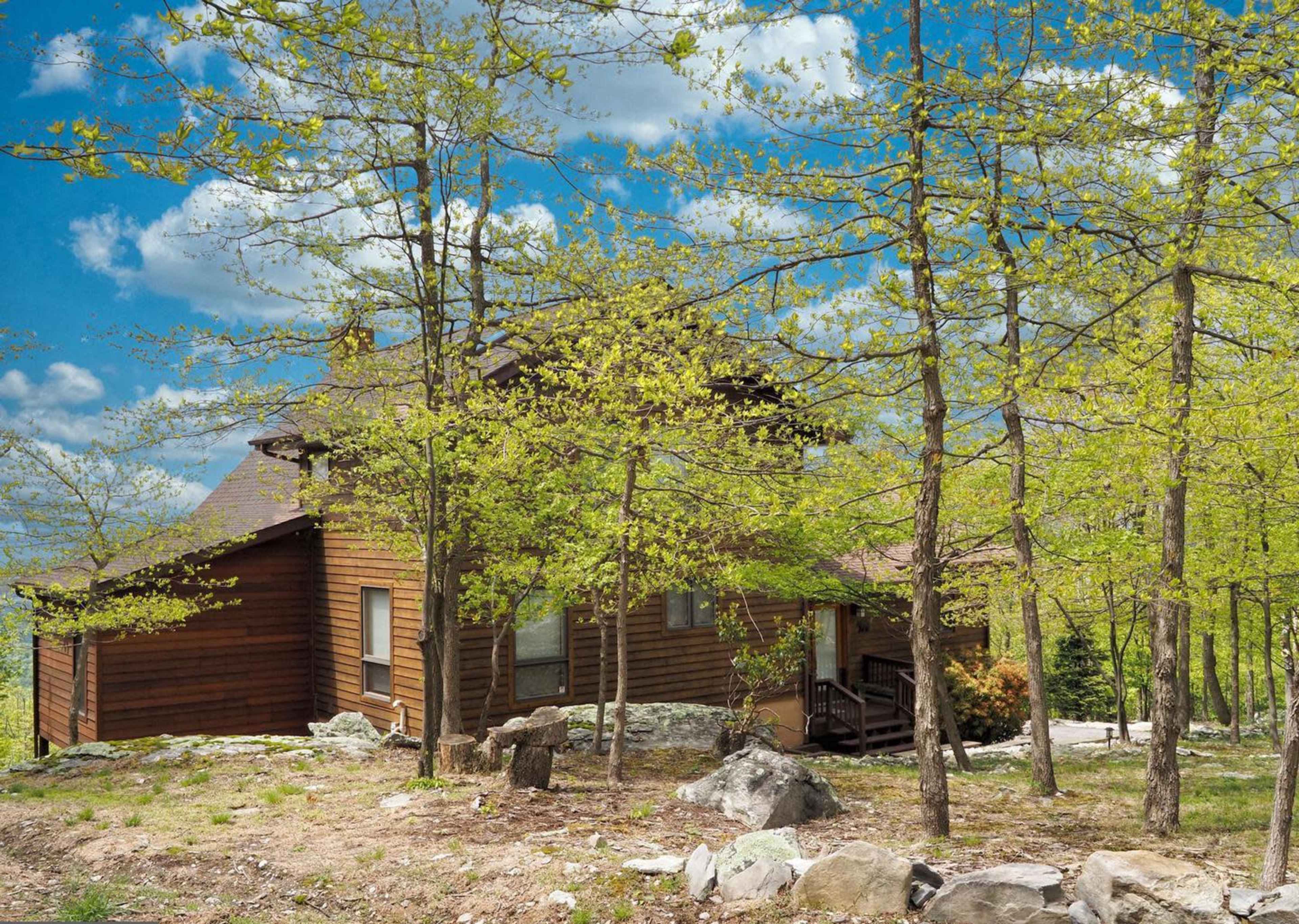 A wooden cabin surrounded by young green trees and large stones under a partly cloudy blue sky.