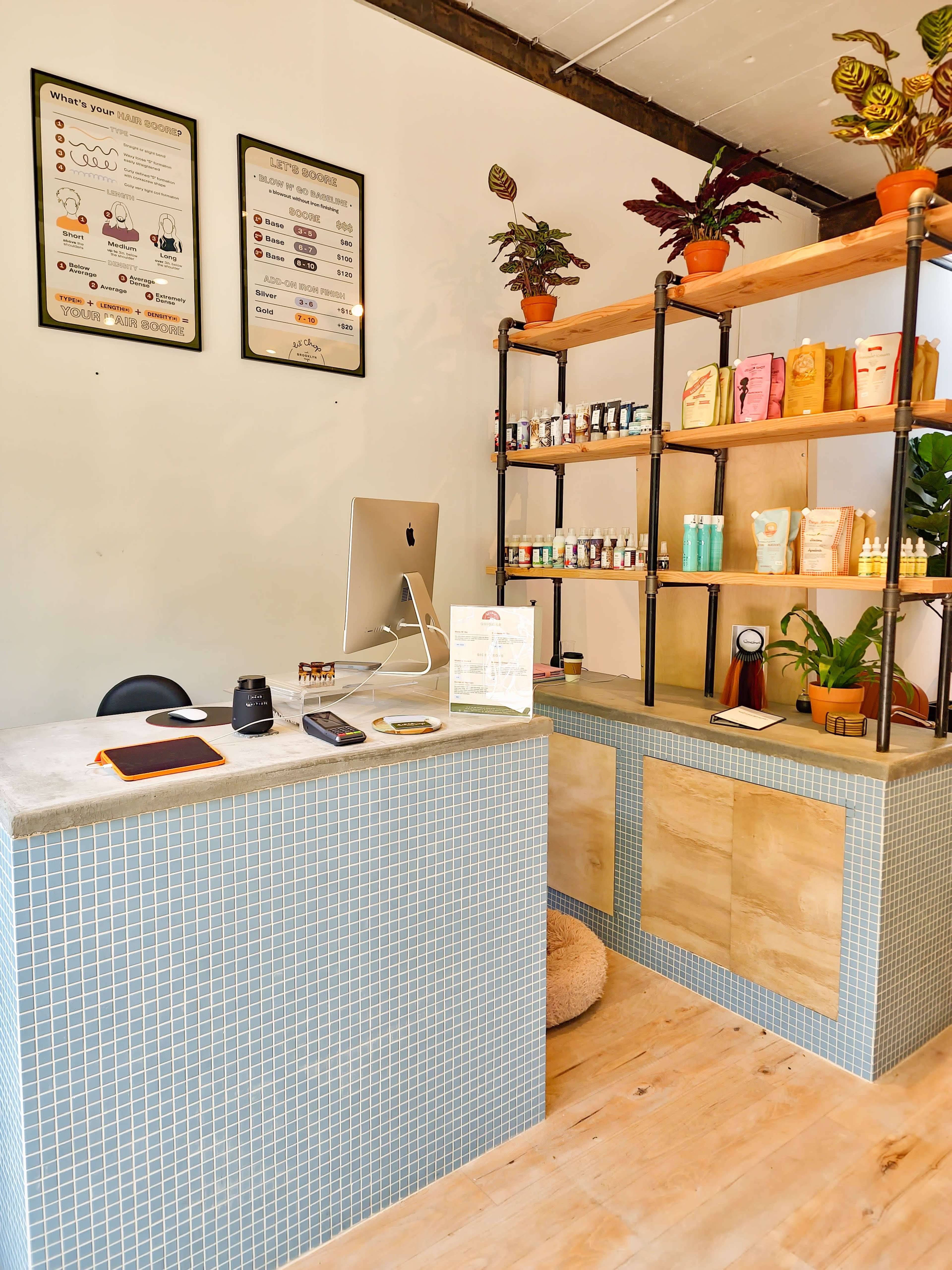 A reception area with a computer on a desk, a display shelf filled with various products, and plants positioned on top of the shelf.