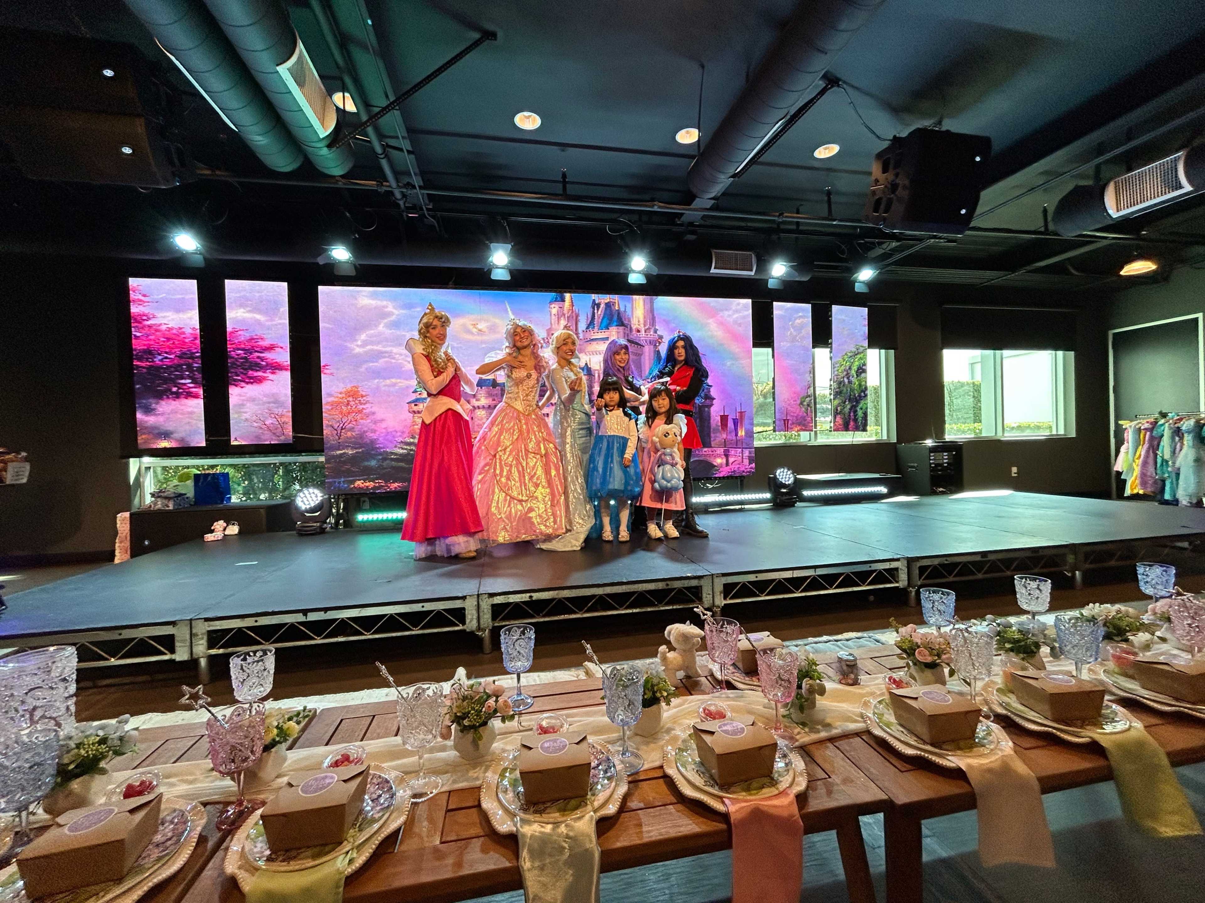 A group of costumed performers and children stands on a stage with a colorful backdrop, while a decorated table is set up in front with small boxes and floral arrangements.