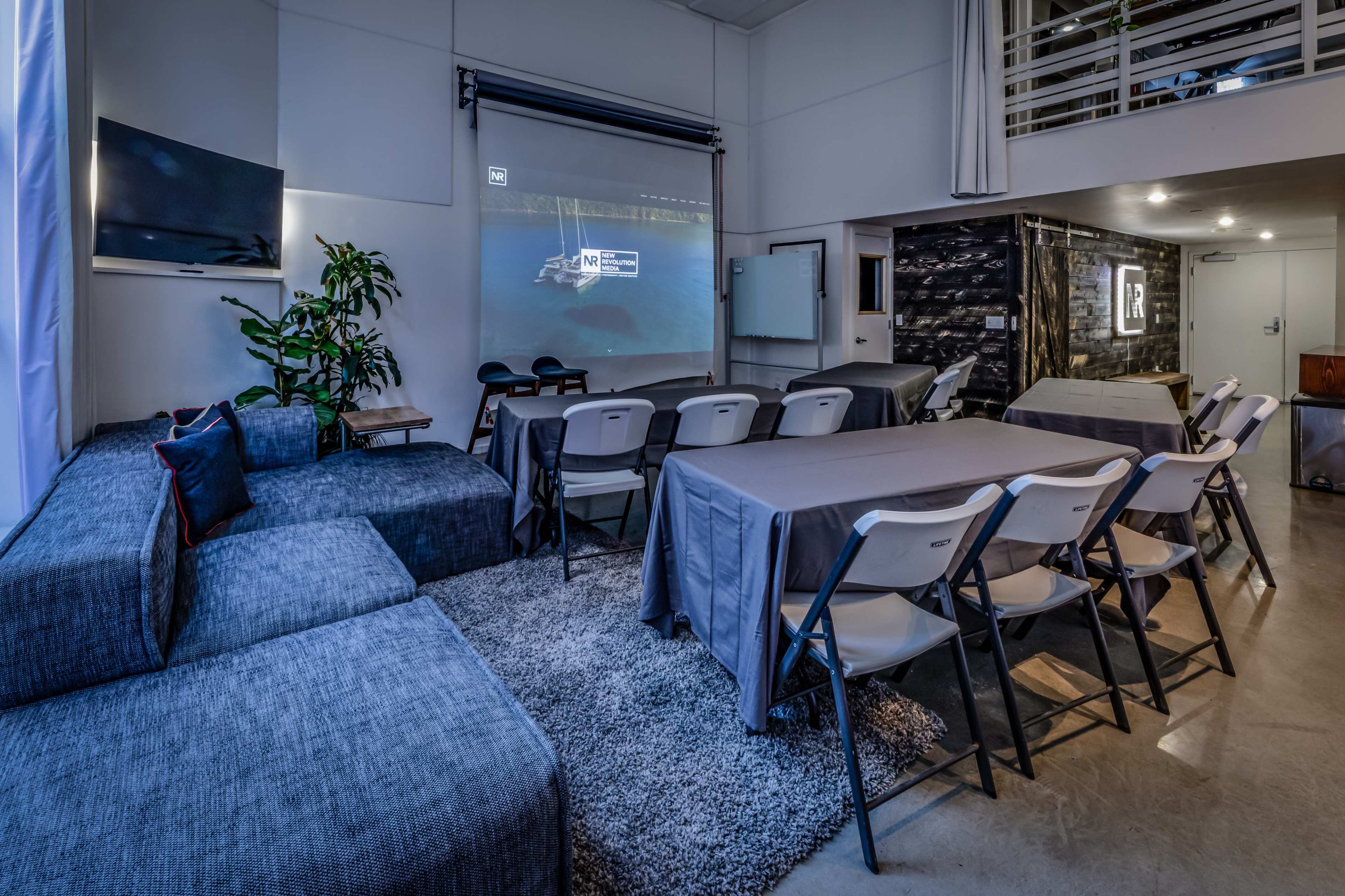 A modern meeting room arranged with several tables and chairs, featuring a large screen for presentations and a comfortable seating area with a sectional sofa.