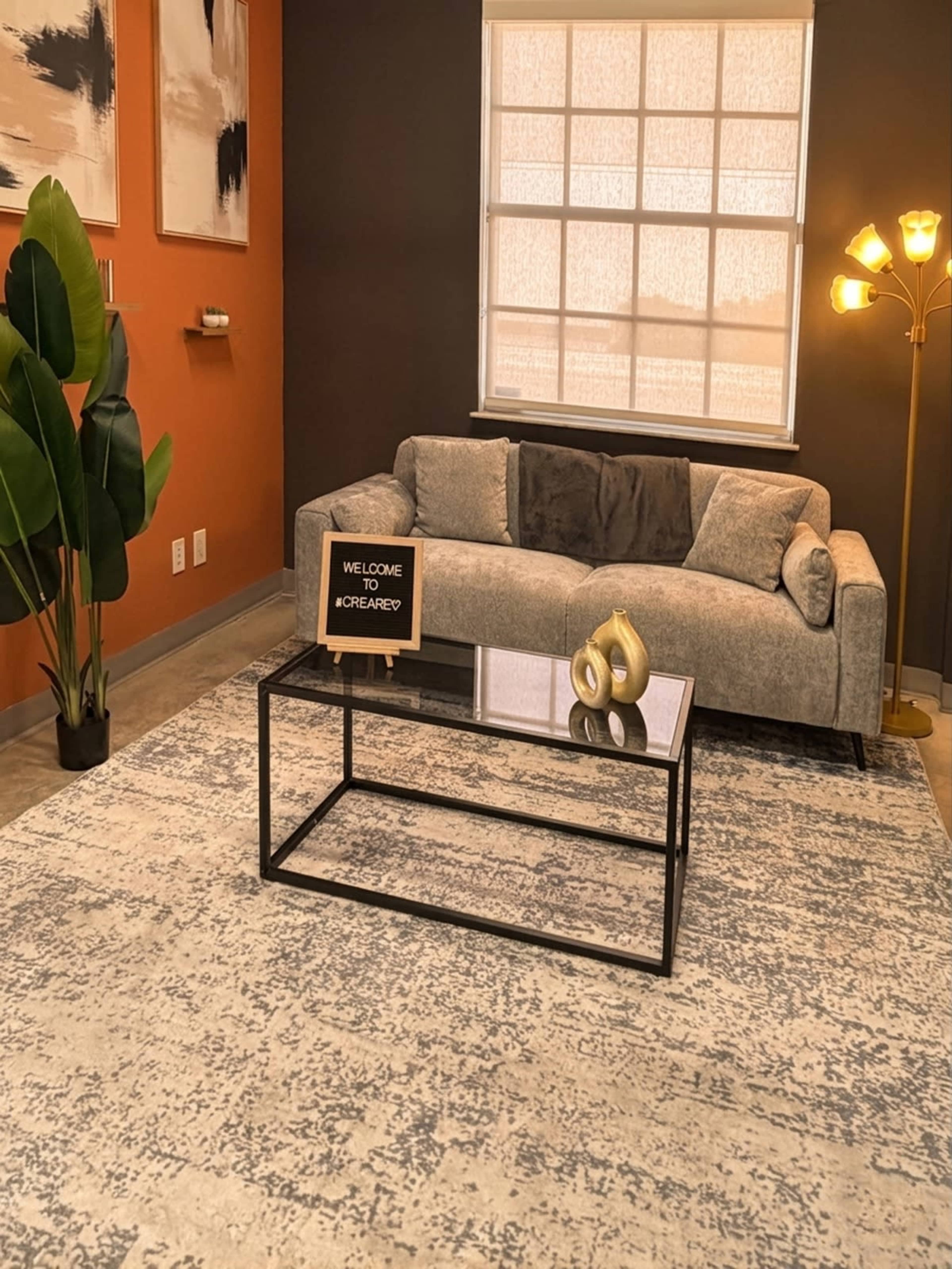 The image shows a modern living room featuring a gray sofa, a glass coffee table with a decorative sign, a plant, and a floor lamp against a backdrop of textured walls.