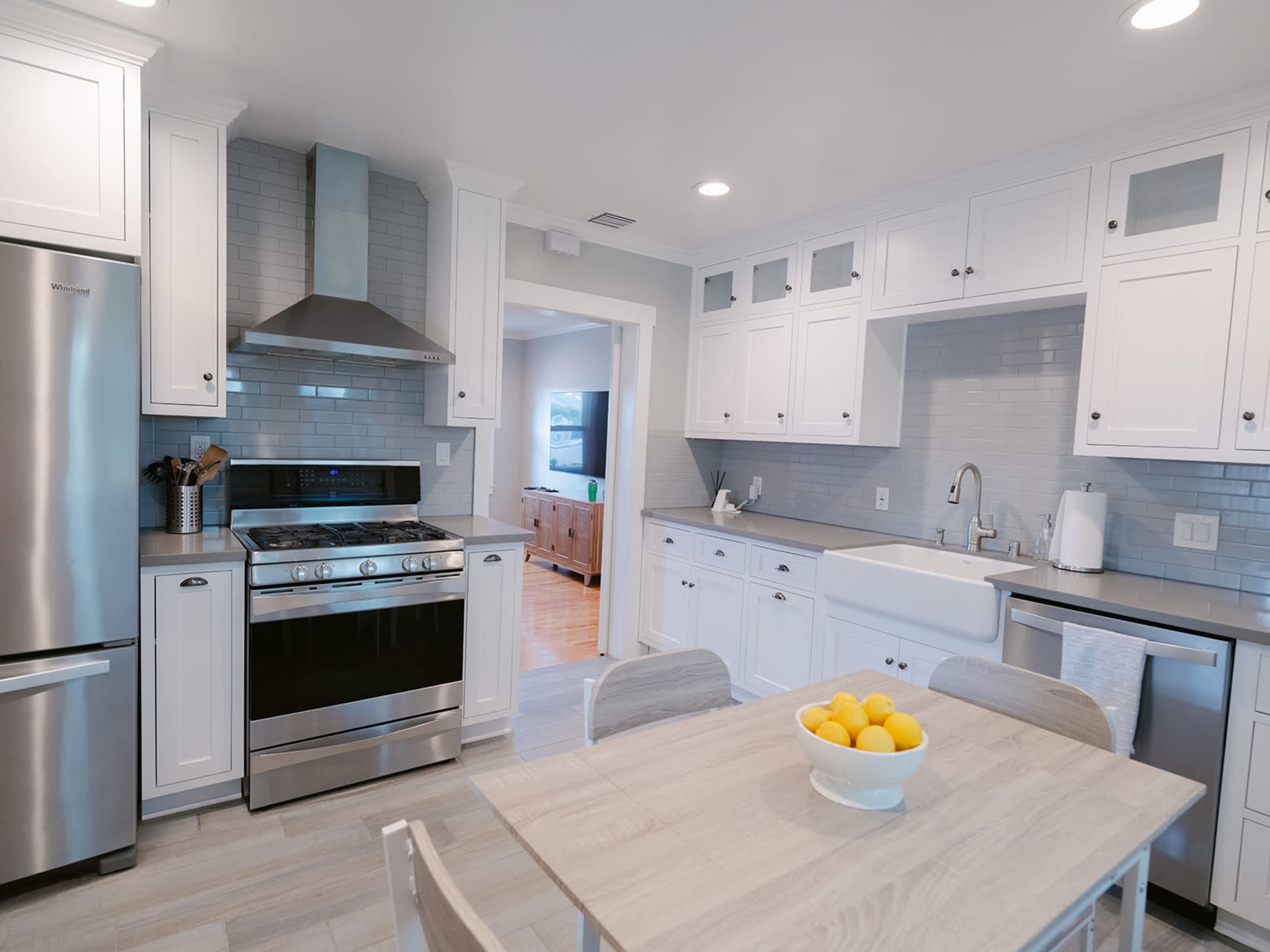 The image depicts a modern kitchen featuring stainless steel appliances, white cabinetry, and a dining table with a bowl of lemons.