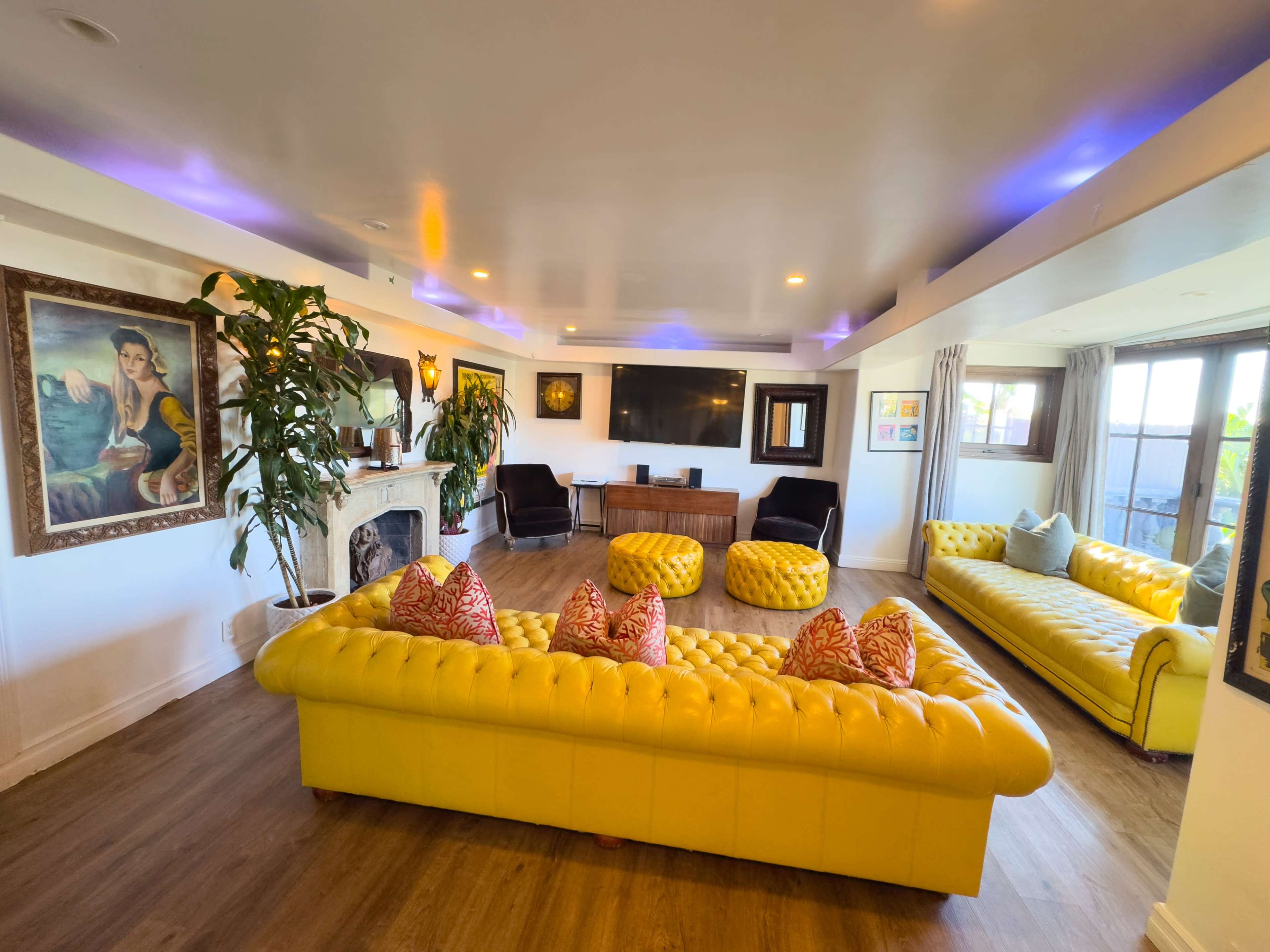 The image shows a brightly lit living room furnished with yellow tufted sofas, several colorful cushions, a large flat-screen TV, and a framed portrait on the wall.