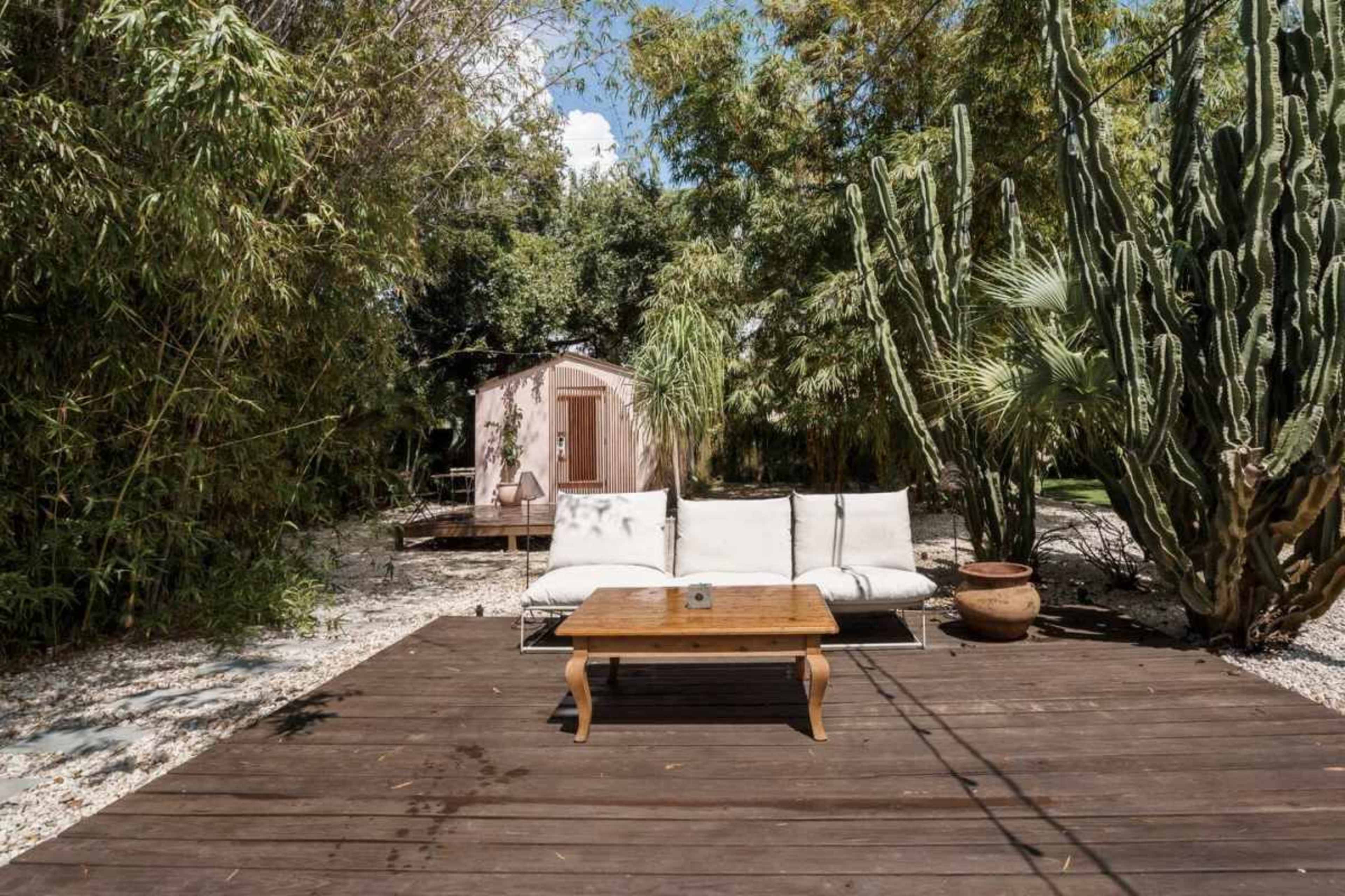 The scene features a wooden deck with a white couch and coffee table surrounded by lush greenery and cacti, with a small shed visible in the background.