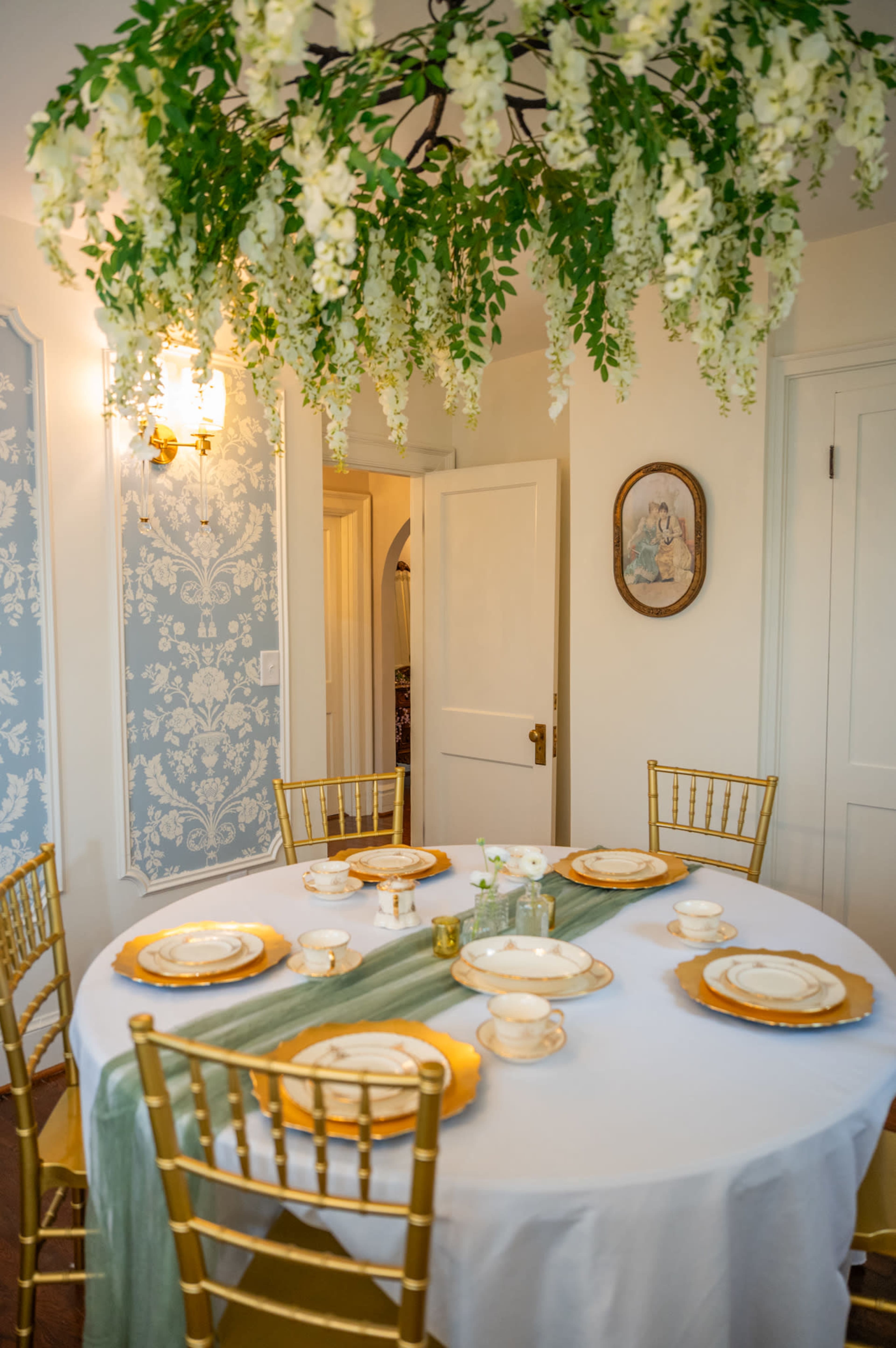 The image shows a dining room with a round table set for six, featuring a white tablecloth, gold-rimmed plates, and a decorative green and gold runner, with floral decorations hanging above.