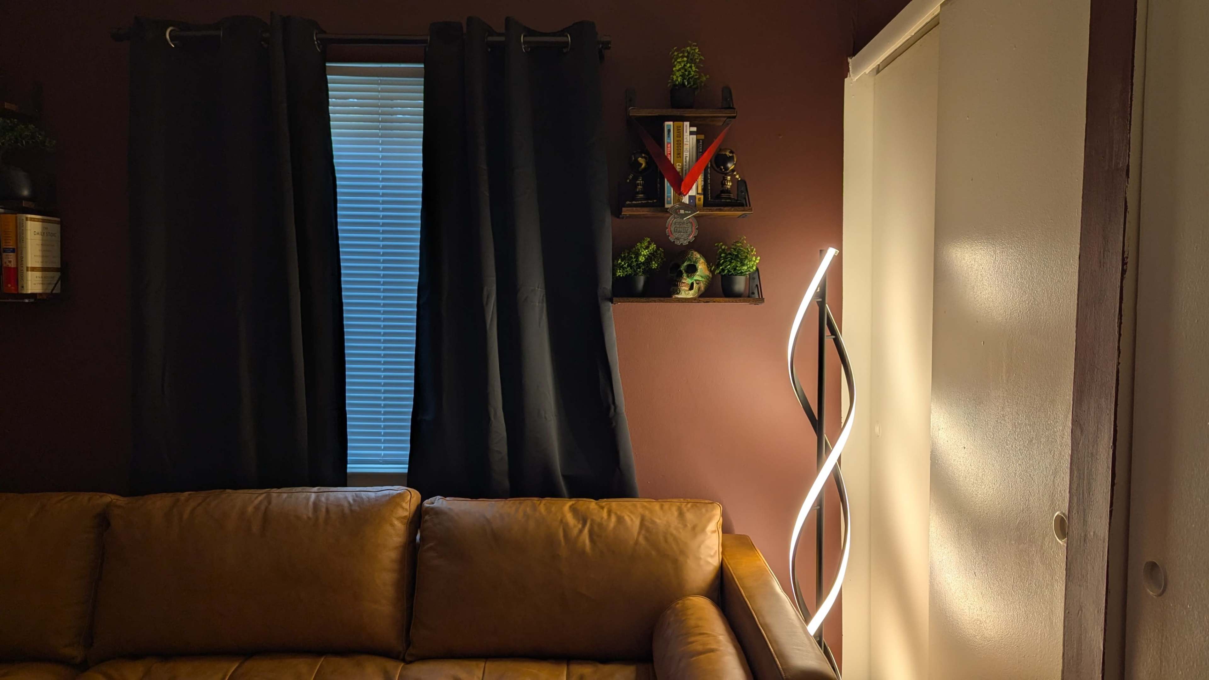 The image shows a cozy living room corner featuring a brown leather couch, a window with black curtains, a shelf with small plants and decorations, and a modern light fixture.