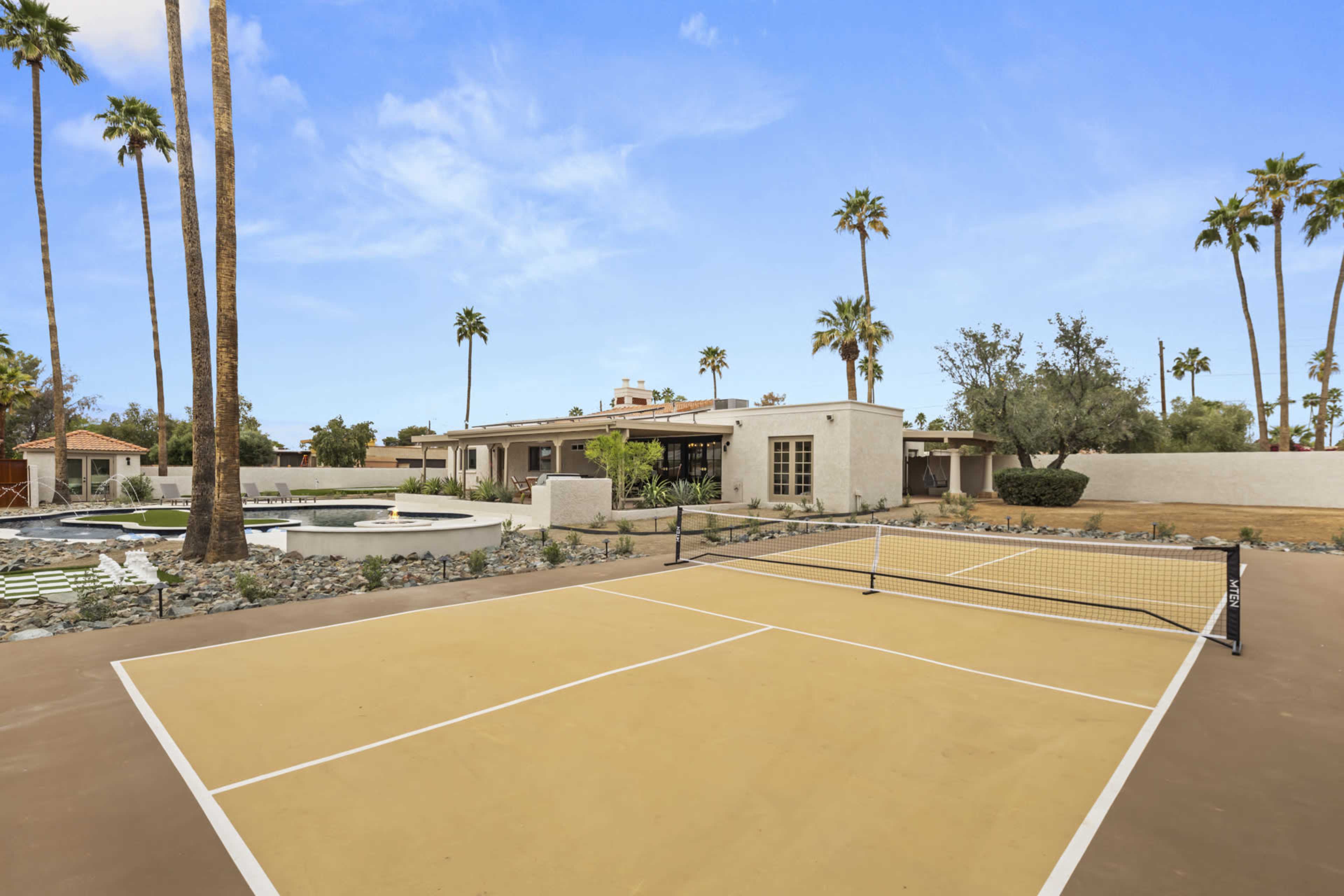The image features a tennis court in the foreground, with a house and palm trees visible in the background.