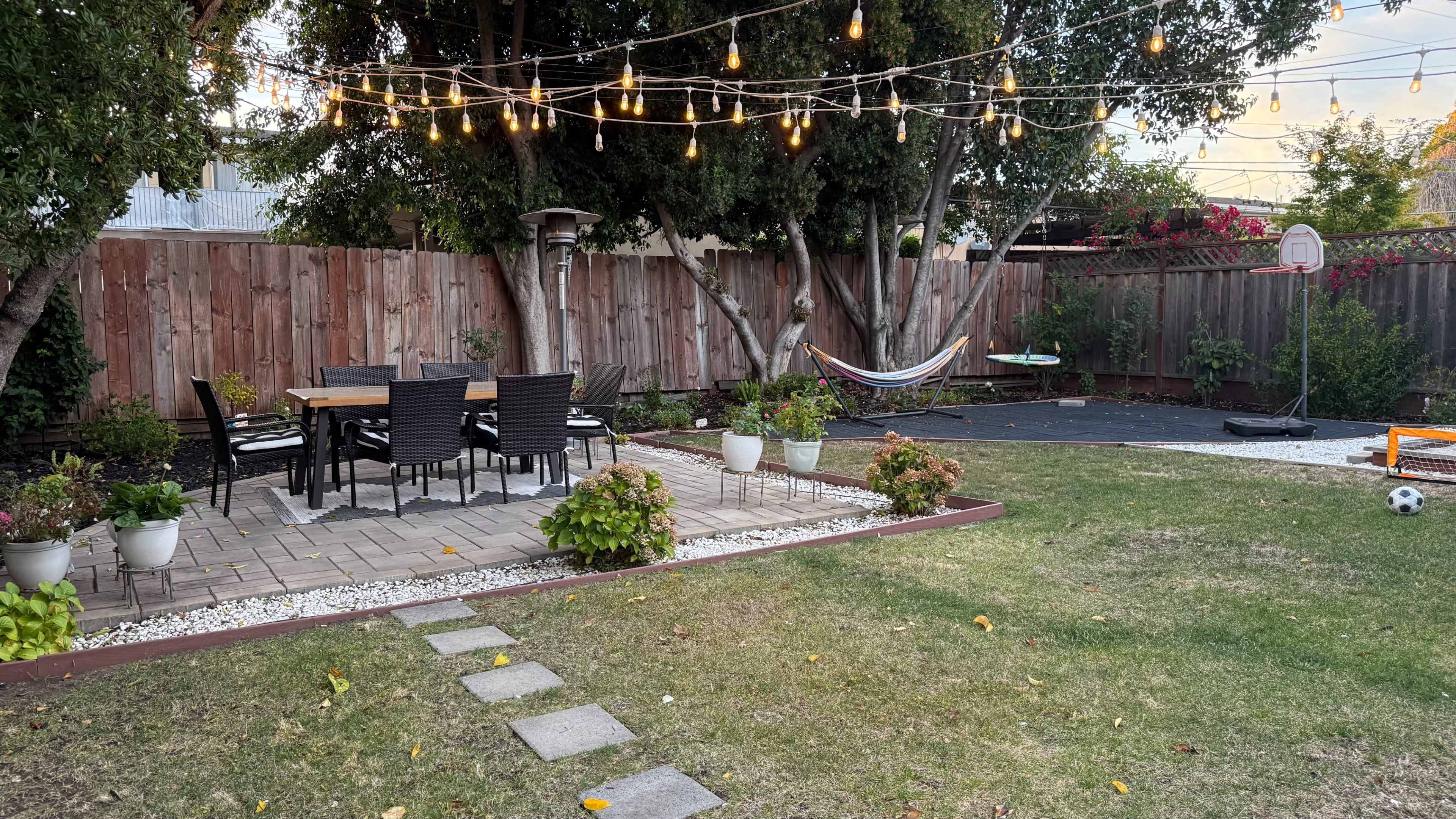 The image shows a backyard with a dining set under string lights, a hammock, a small paved area, and a grassy section alongside potted plants.