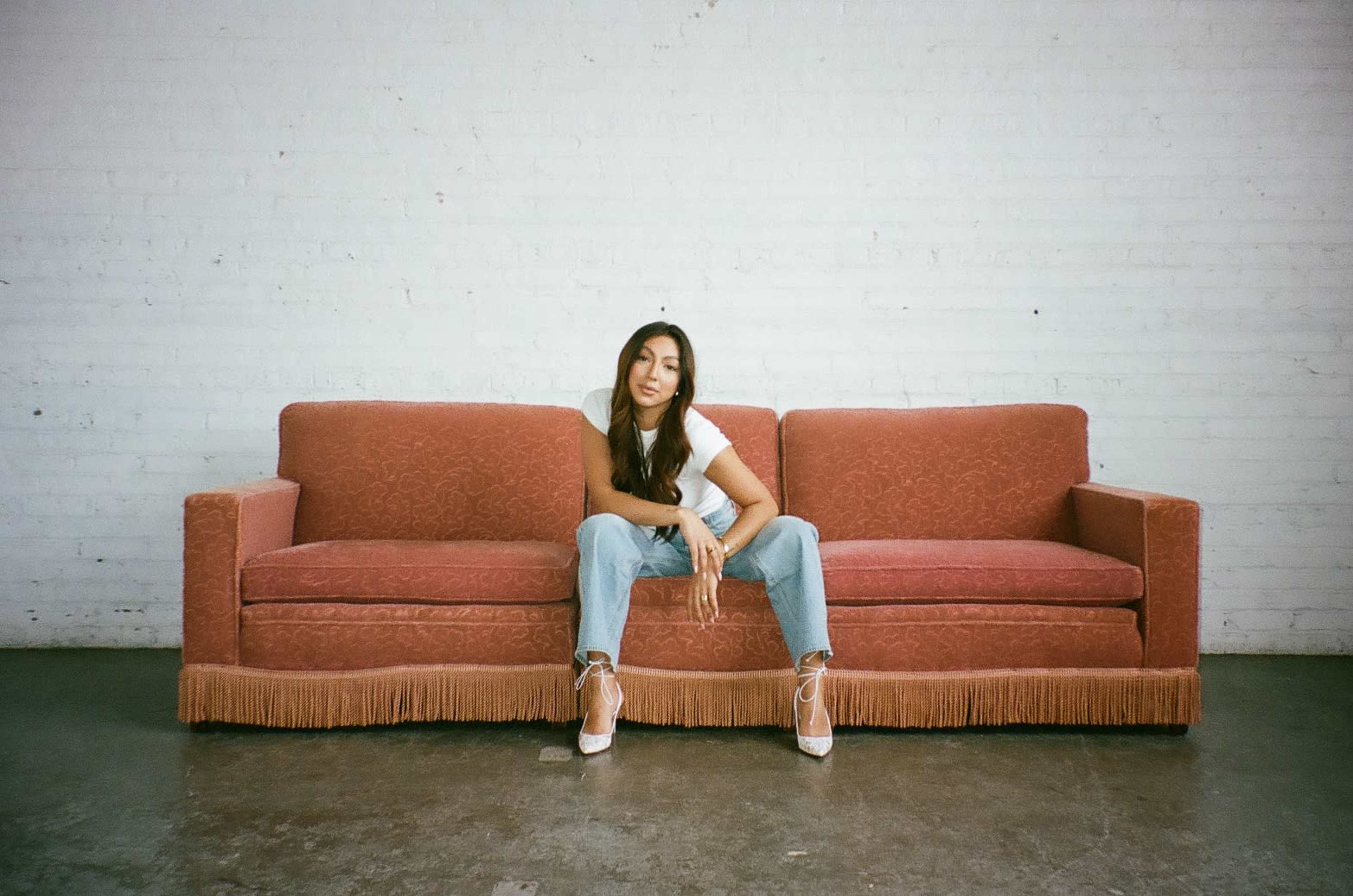 A woman sits on a vintage red couch against a white brick wall.