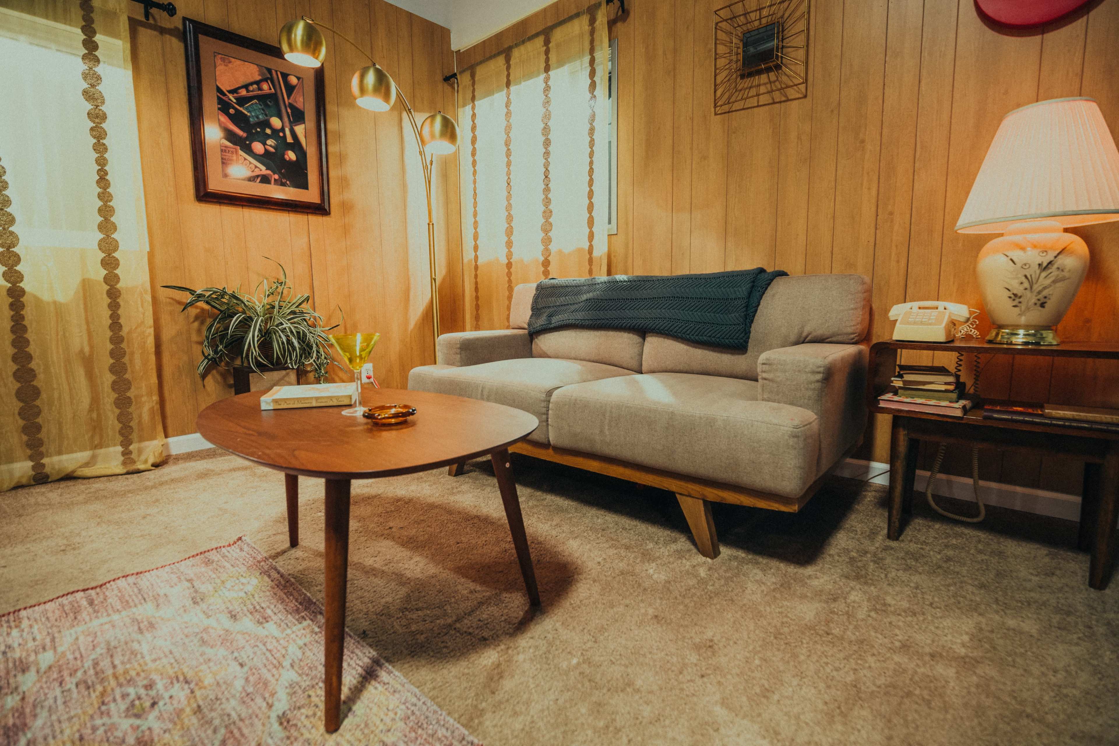 A cozy living room with a gray sofa, a wooden coffee table, a lamp, and decorative curtains against a paneled wall.
