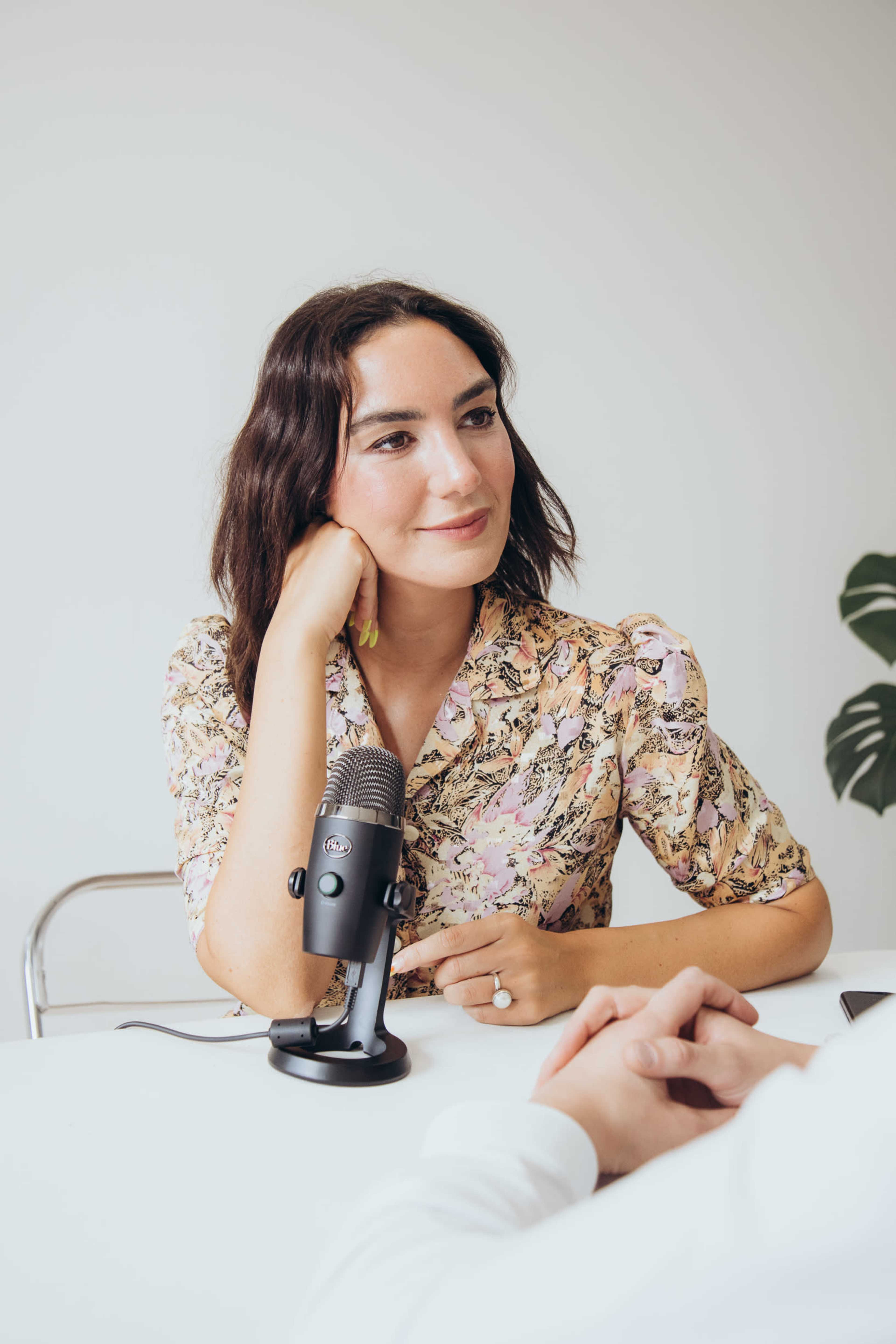 A woman in a floral dress sits at a table with a microphone, engaged in conversation.
