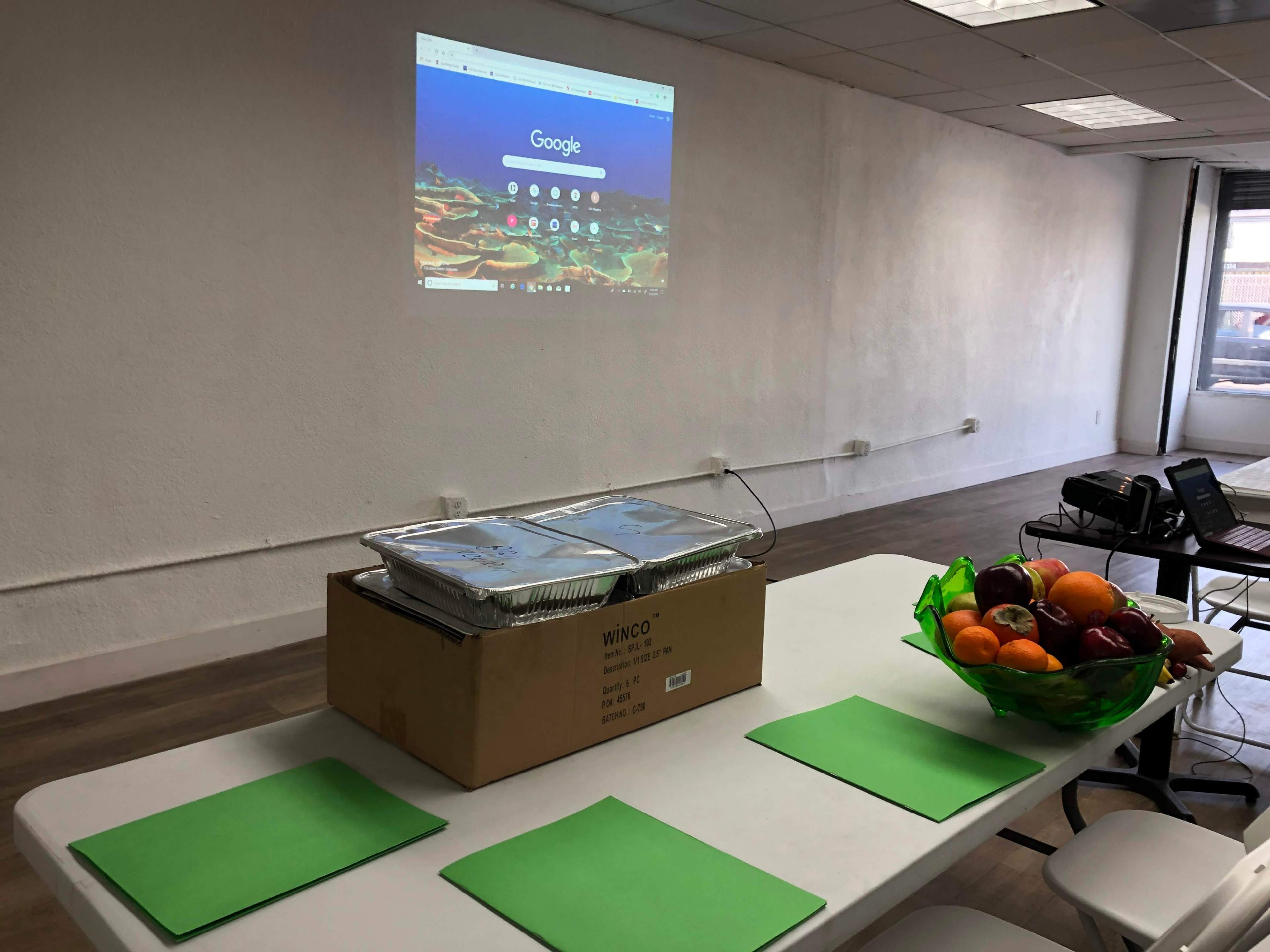 A table is set up with food containers and a bowl of fruit, while a projector displays a Google homepage on the wall in an empty room.