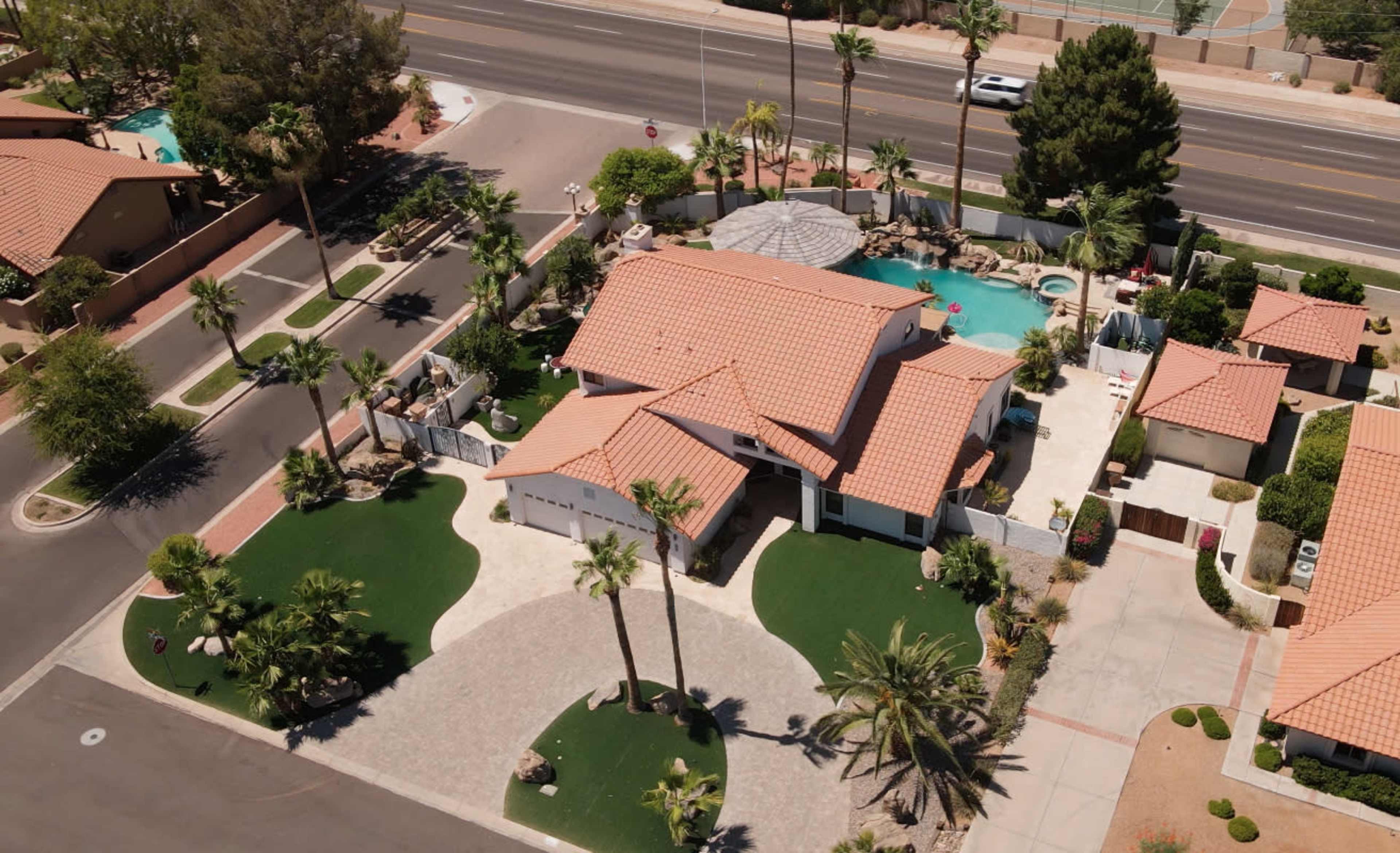 An aerial view of a suburban home surrounded by neatly landscaped lawns and palm trees, with a pool in the backyard and a road nearby.