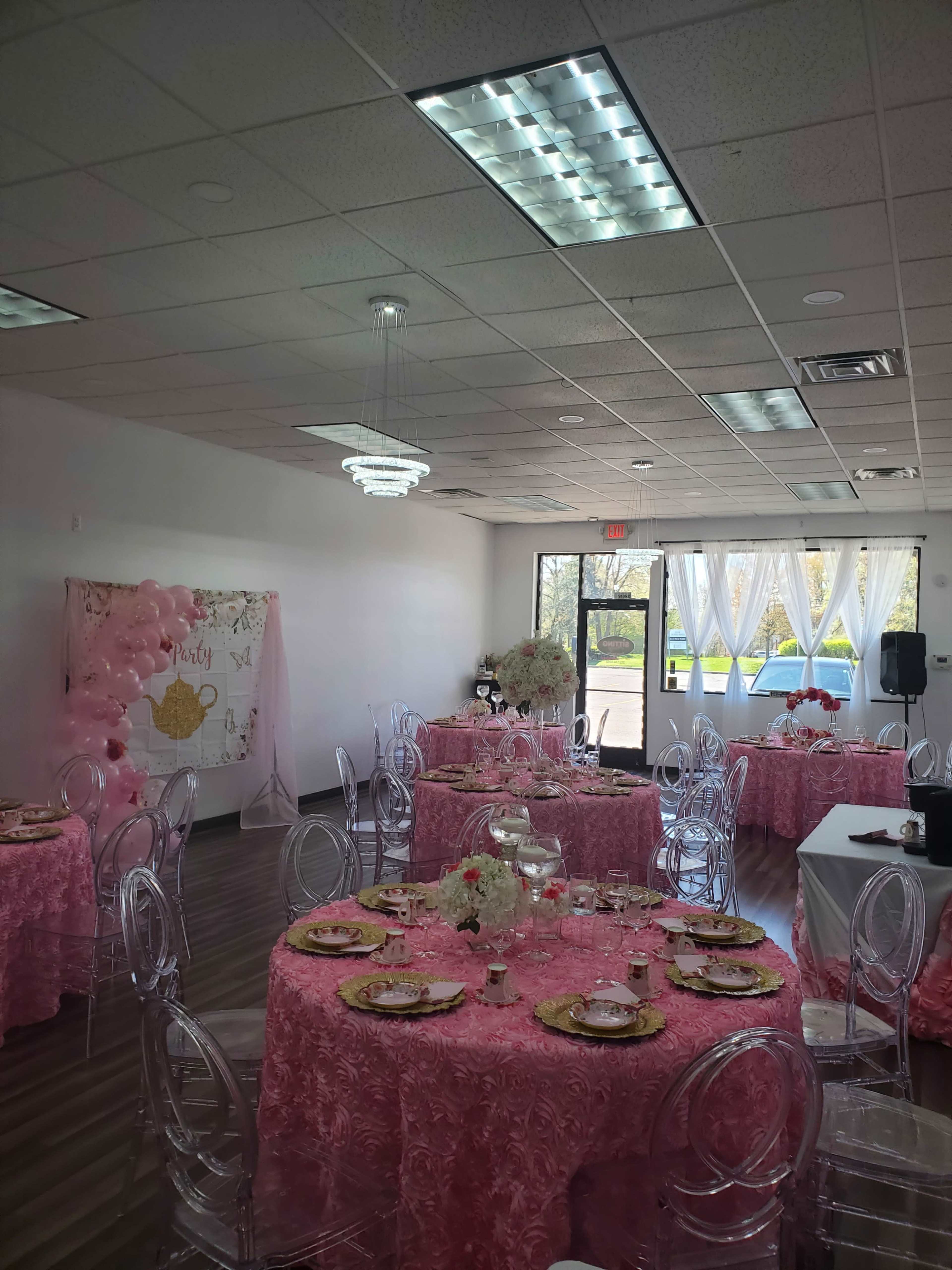 A brightly lit event space decorated with pink tablecloths, clear chairs, and floral centerpieces, set up for a formal gathering.