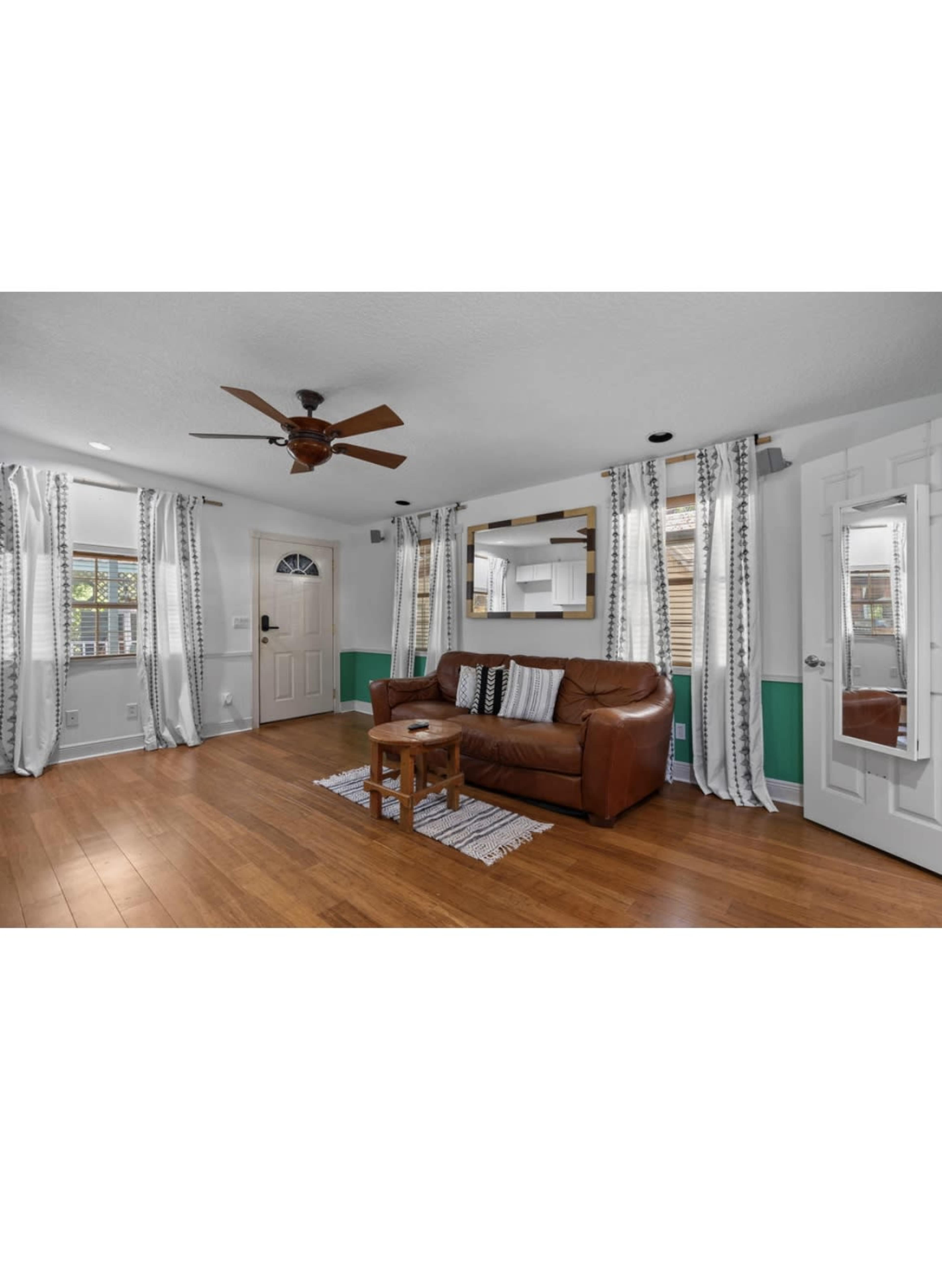 The image shows a living room with a brown leather sofa, a small wooden table, and large windows dressed with striped curtains.
