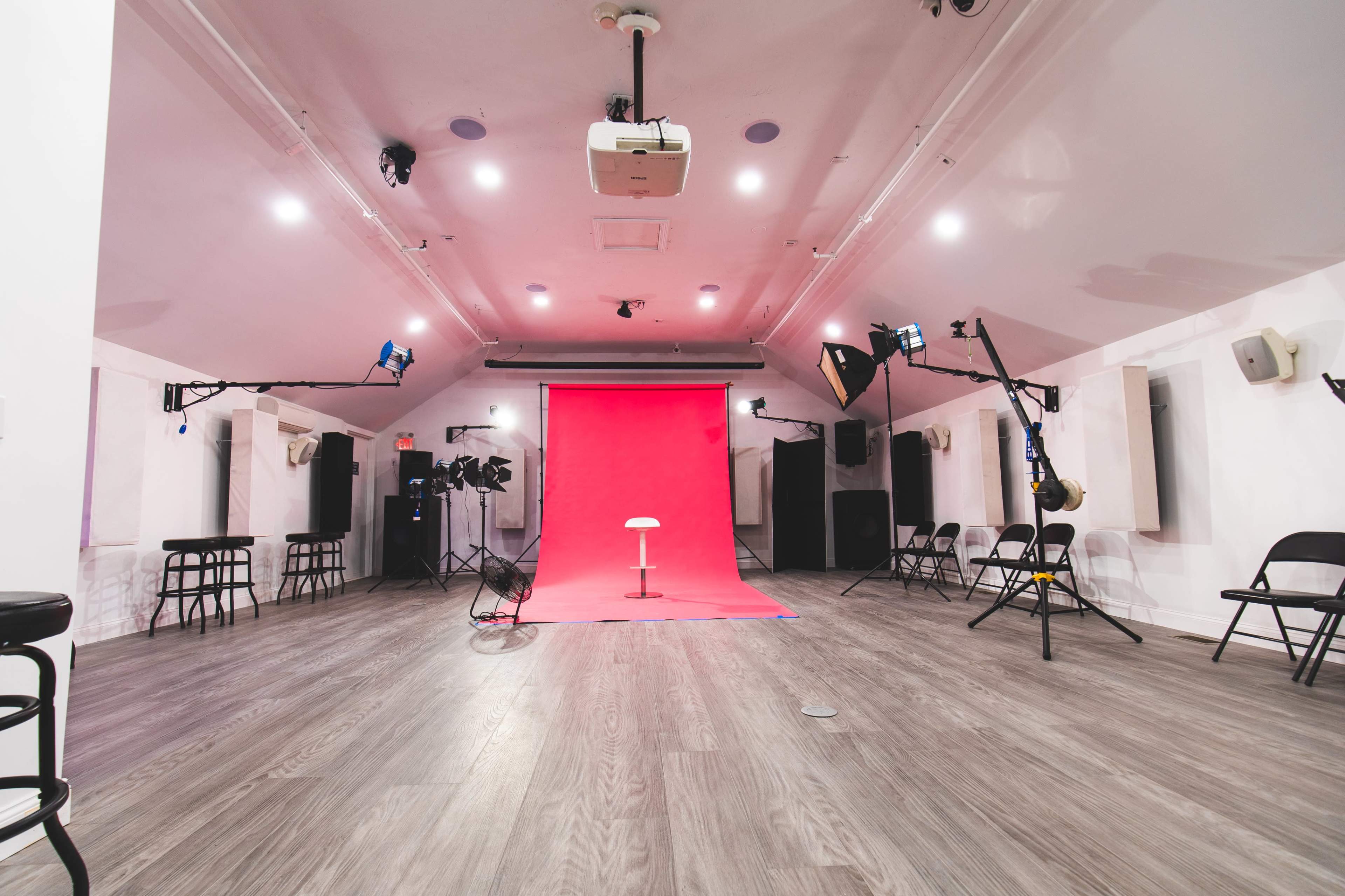 The image shows a studio space with a pink backdrop, lighting equipment, and chairs arranged around the perimeter.