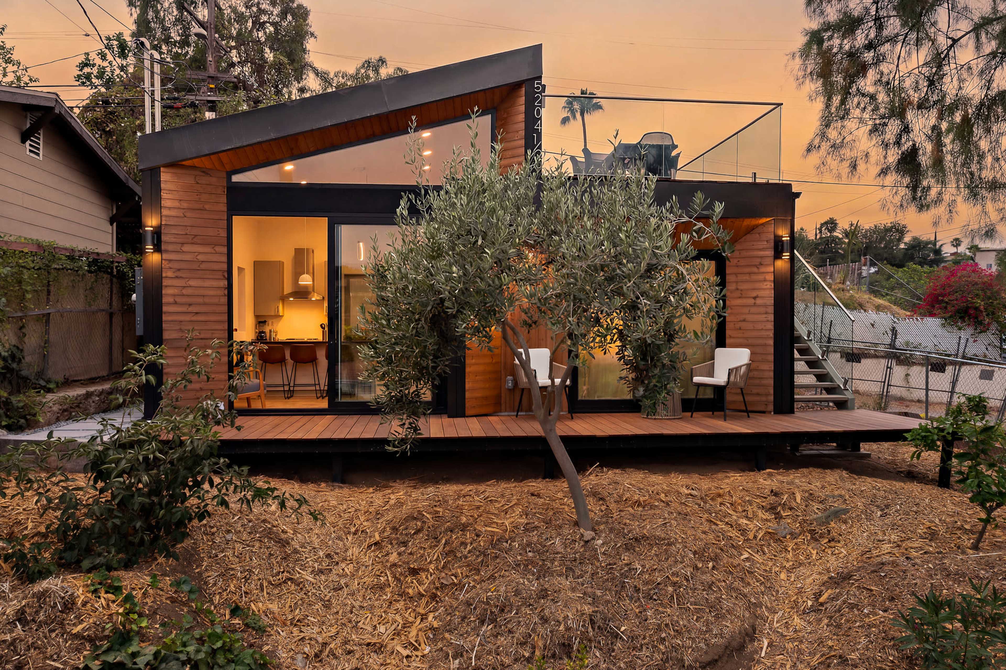 The image shows a modern house with a wooden exterior, large windows, and a landscaped area featuring olive trees and a deck.