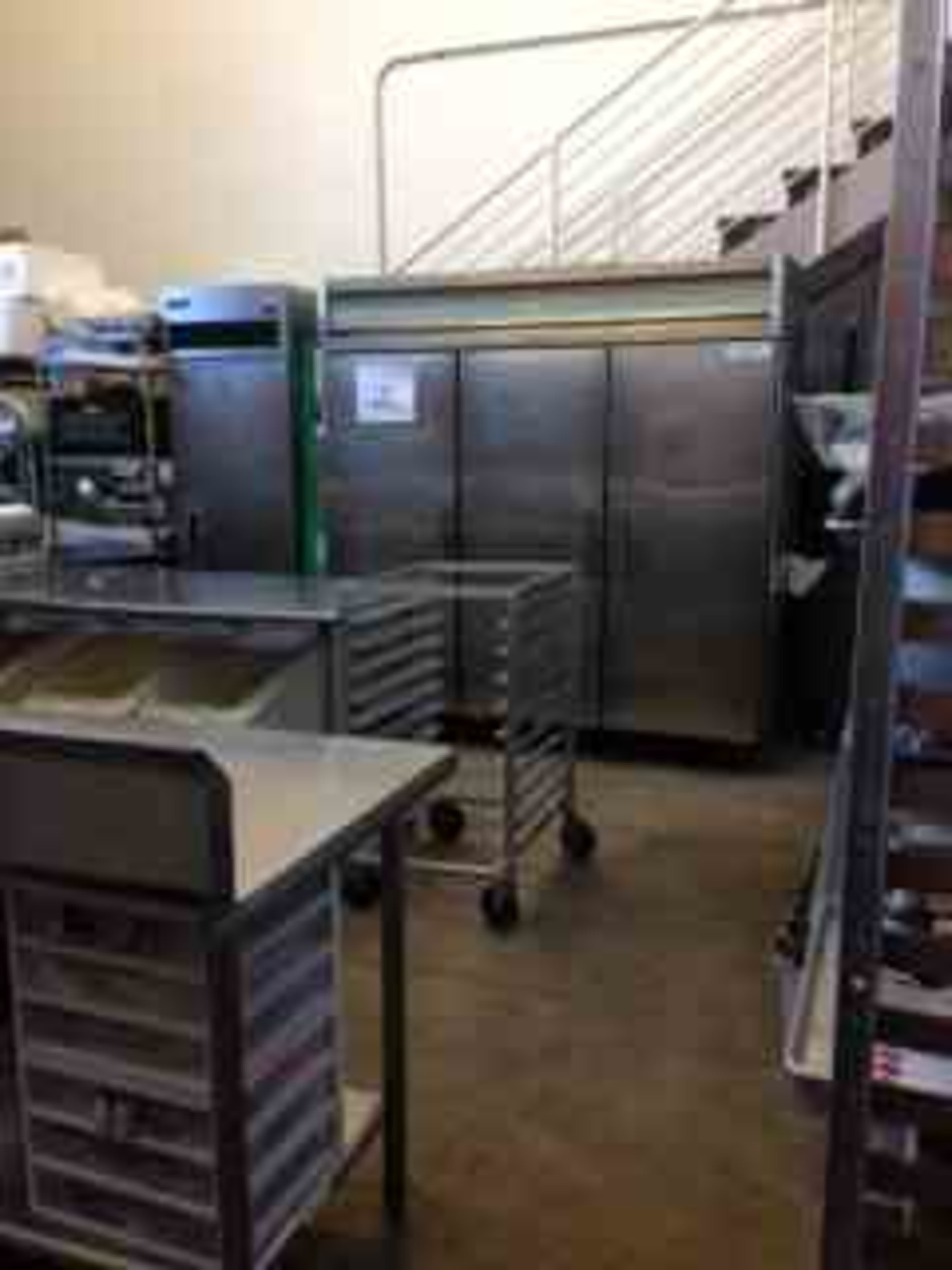 A commercial kitchen area features stainless steel appliances, including refrigerators and storage carts, with stairs visible in the background.