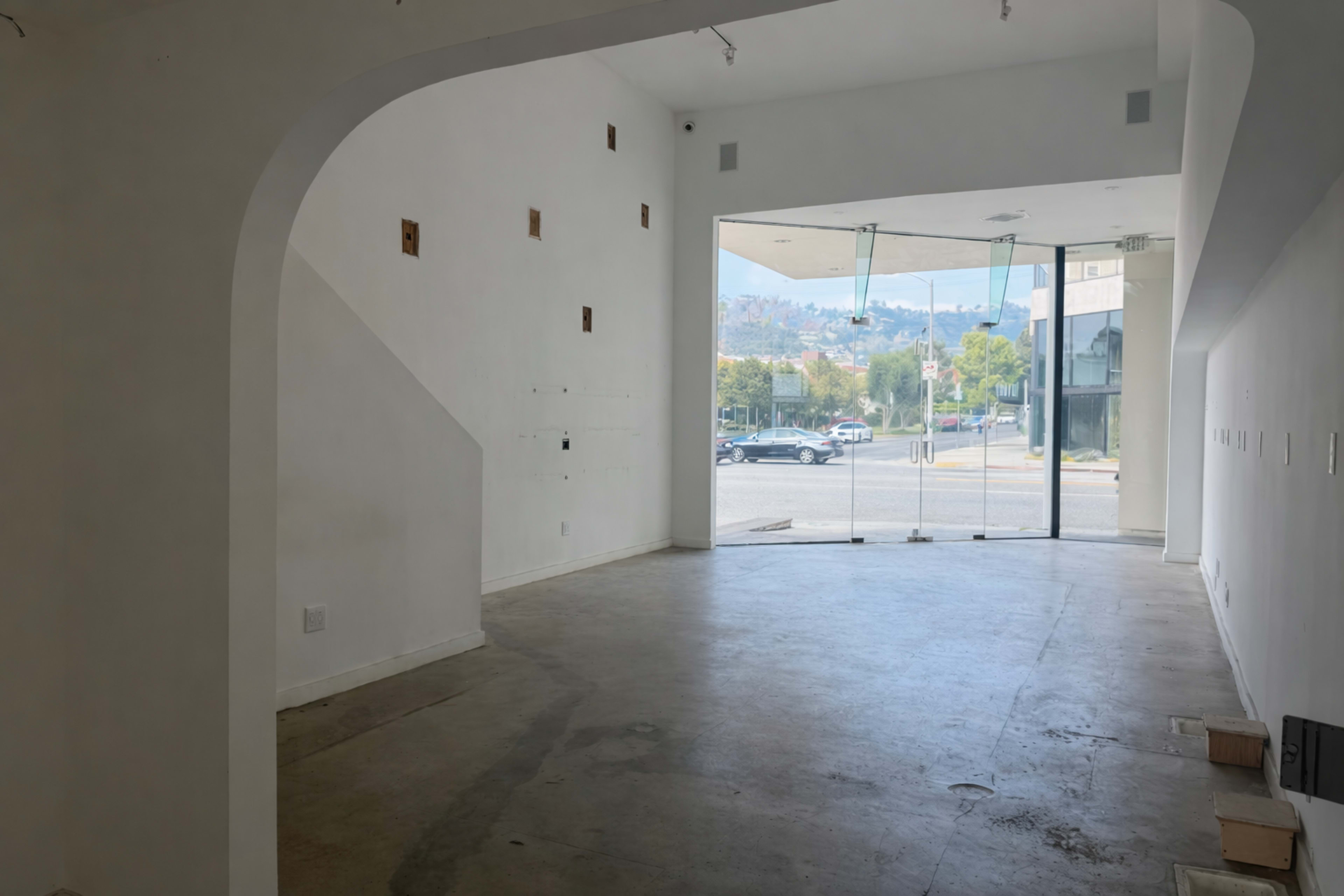 The image shows a spacious, empty interior with a large glass entrance facing a street and minimal wall decorations.