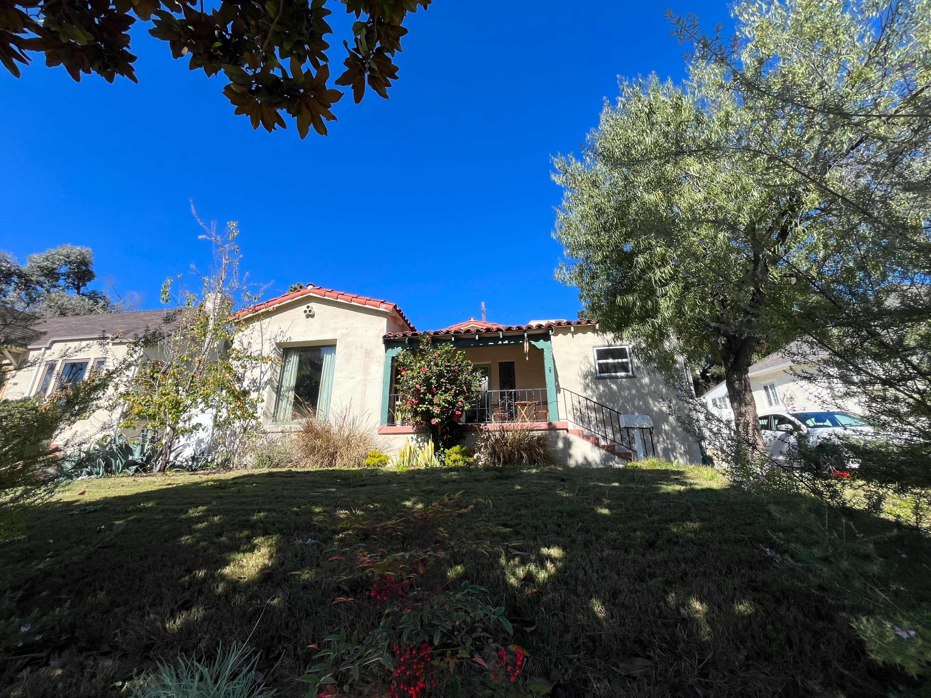 A single-story house with a red-tiled roof is surrounded by greenery under a clear blue sky.