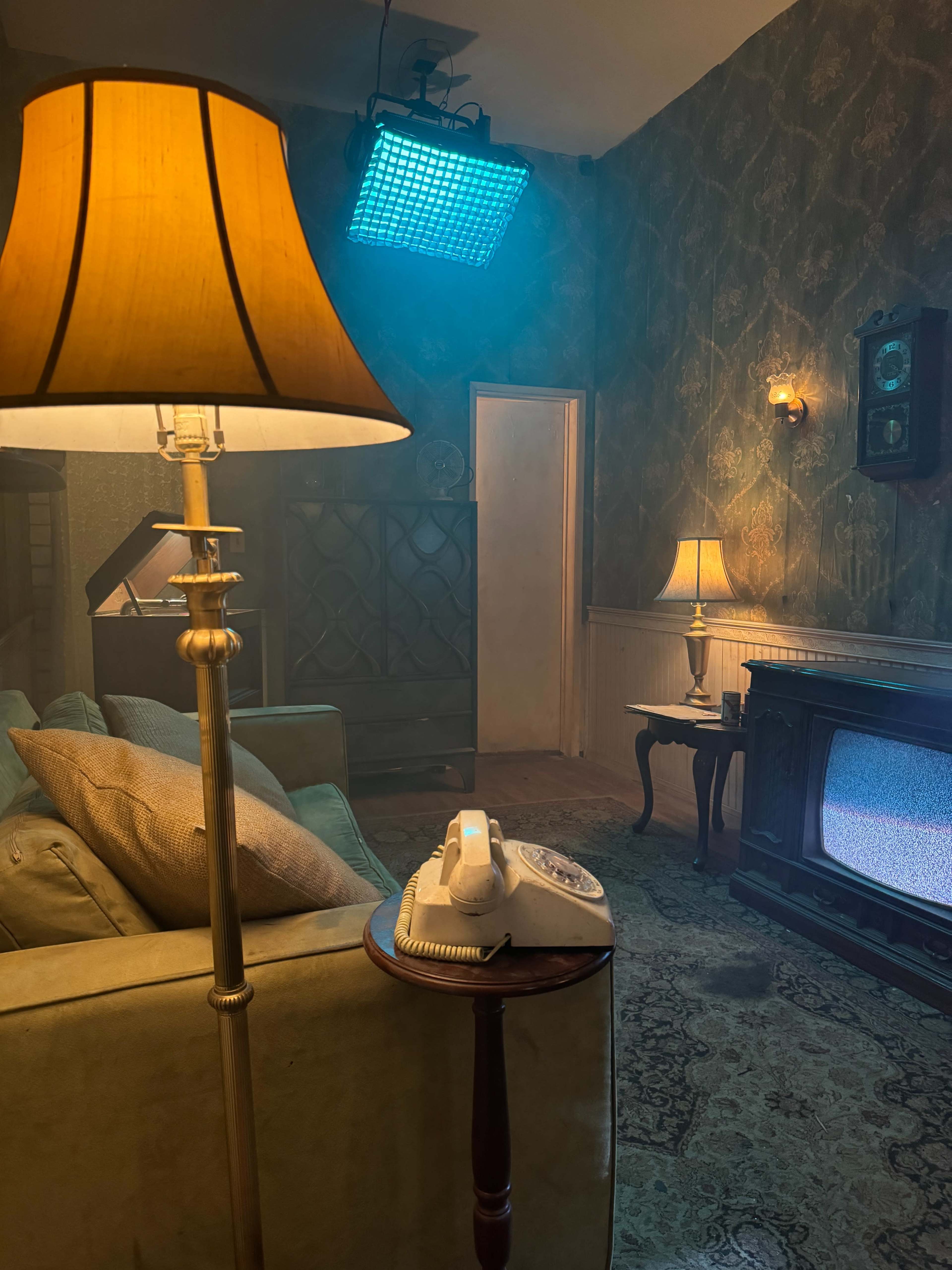 The scene features a dimly lit living room with a vintage telephone on a small table, a green couch, a glowing lamp, and a television displaying static.