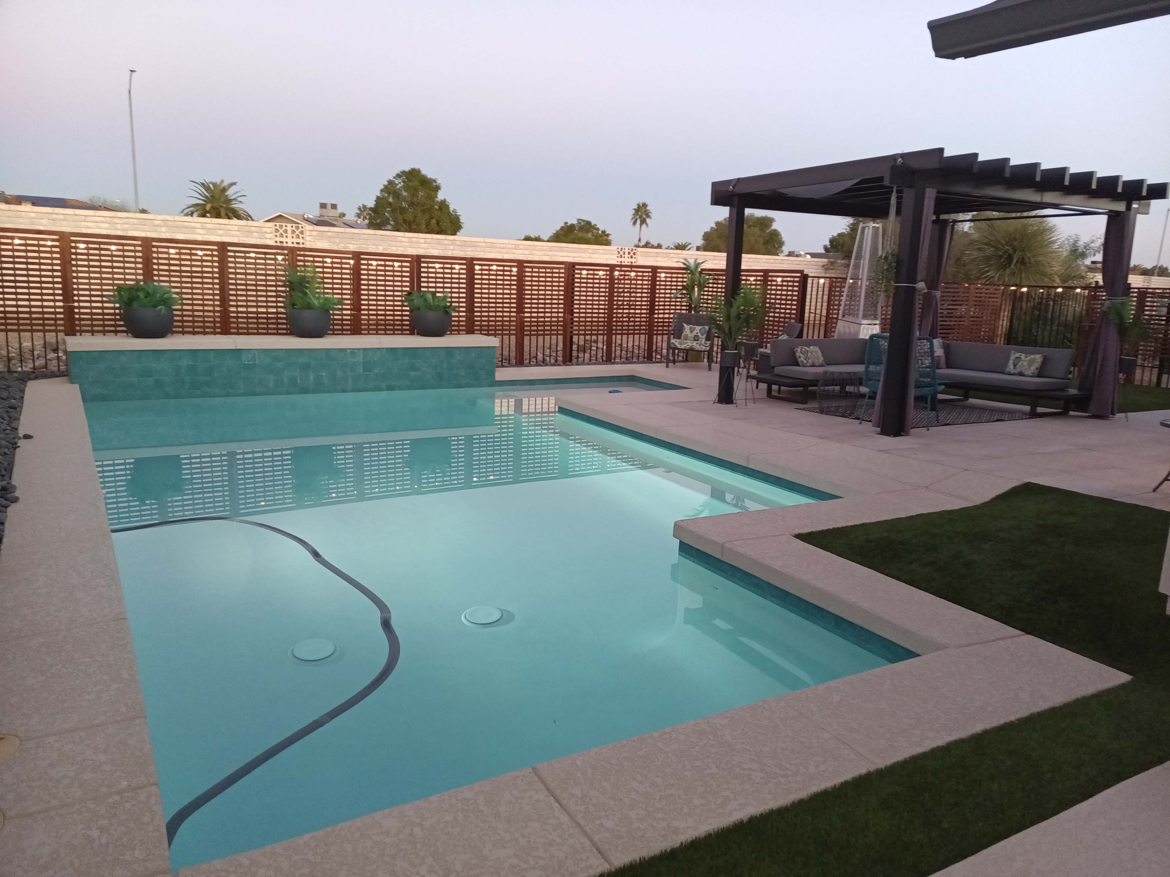 A modern backyard features a swimming pool, a shaded seating area, and landscaped greenery.