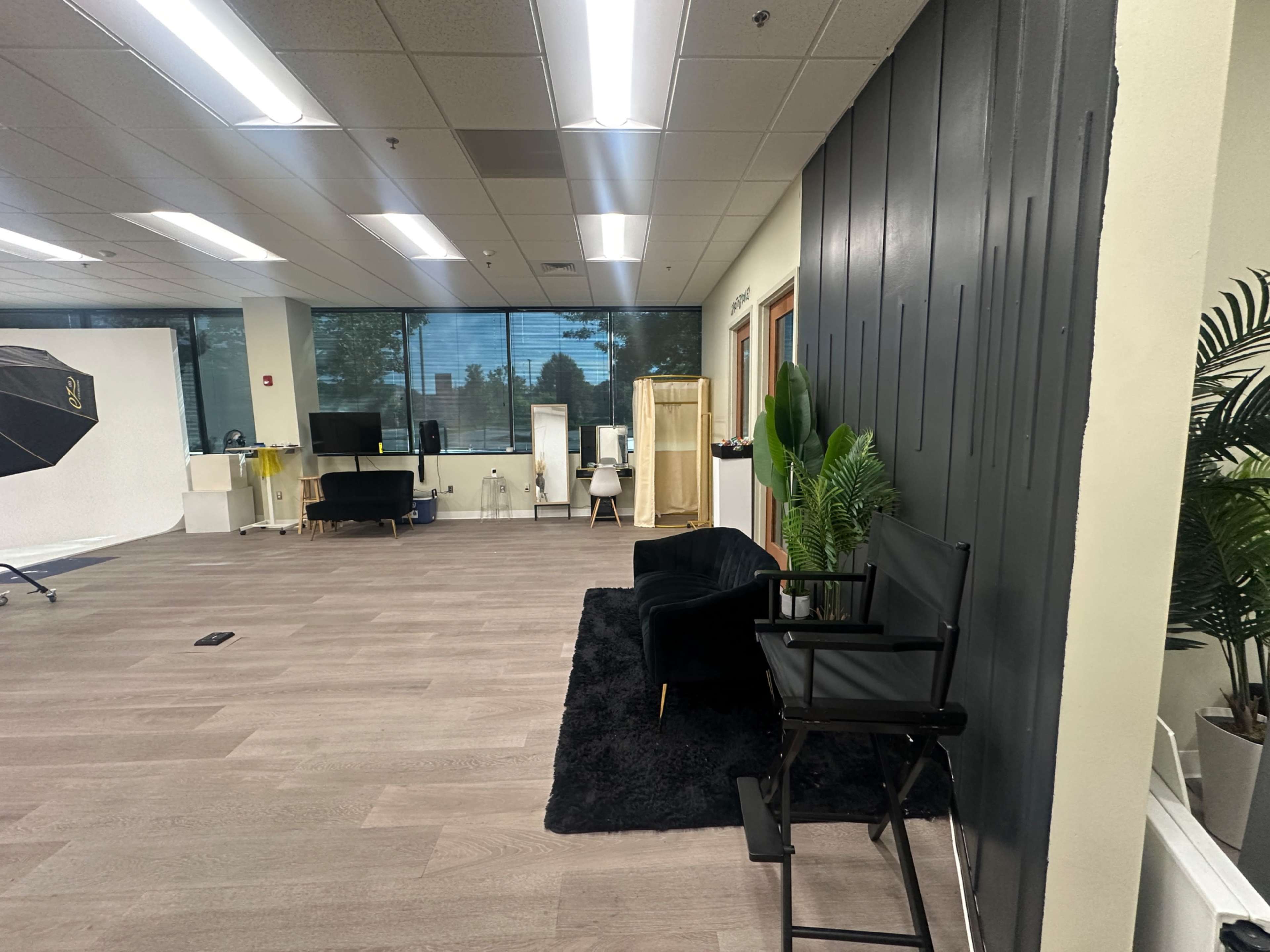 The image shows a spacious room with wooden flooring, featuring a black couch, a director's chair, and large windows that allow natural light to enter.