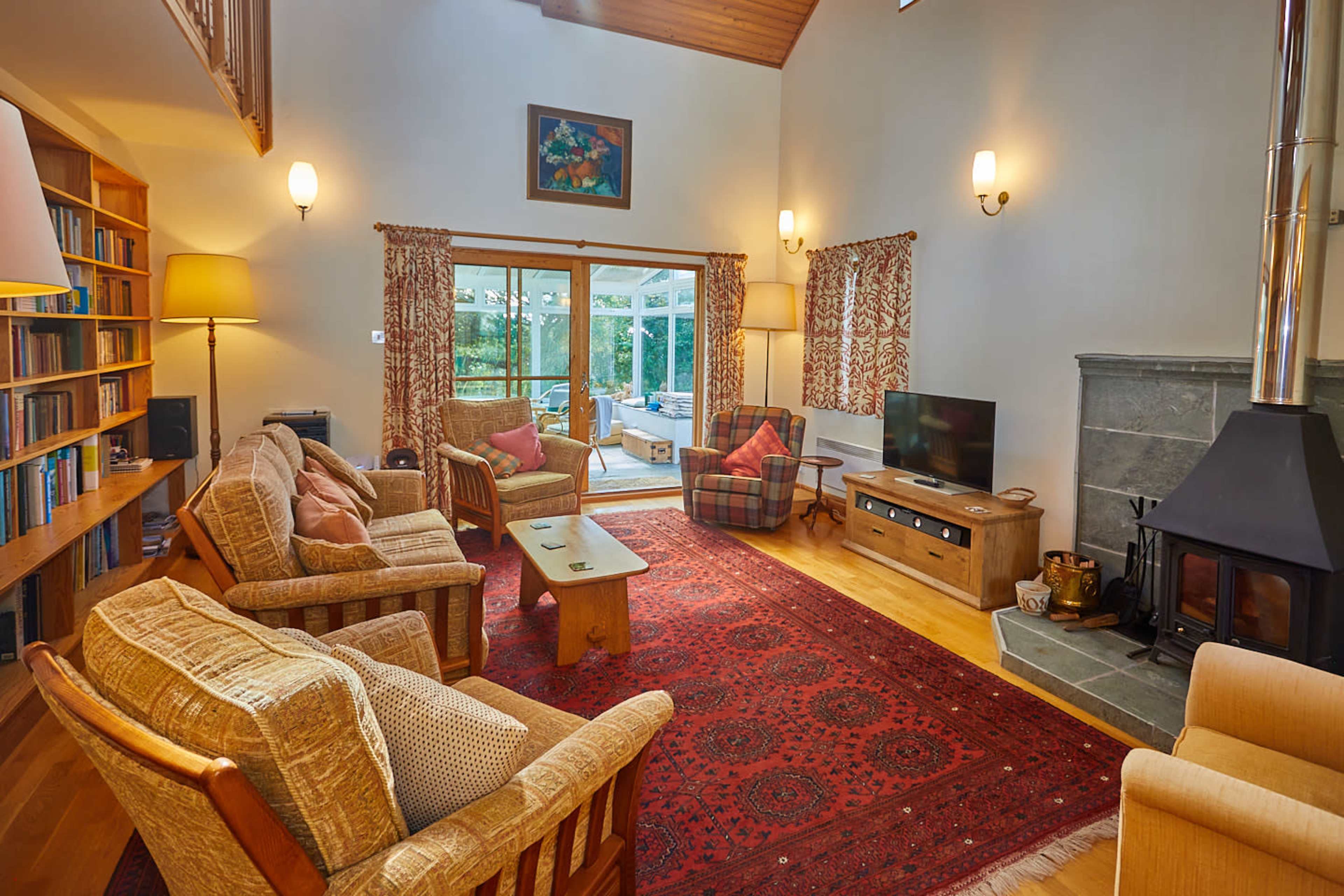 A cozy living room with two sofas, a coffee table, a bookshelf, and a fireplace, featuring large windows that offer a view of the outdoor area.
