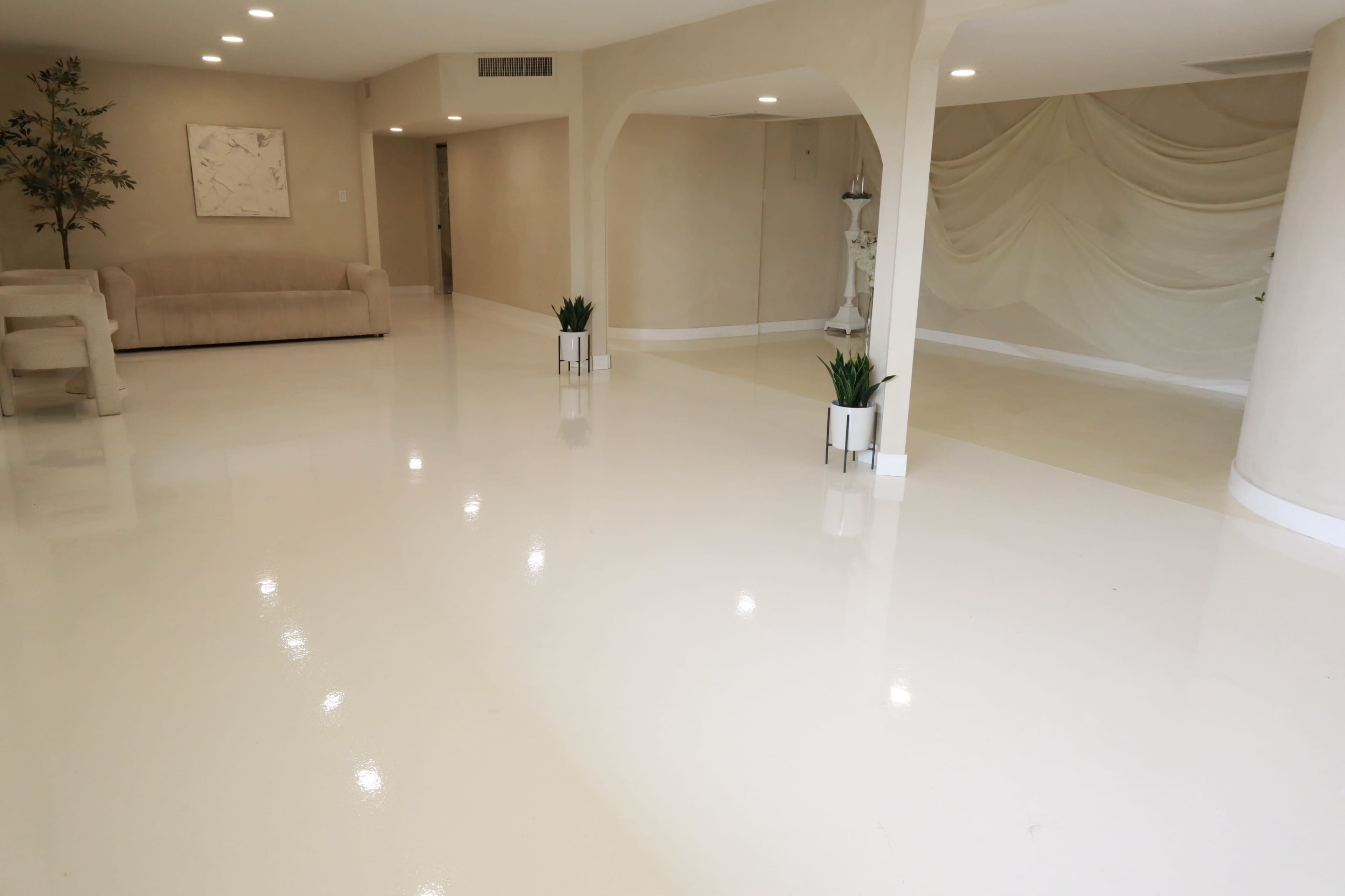 The image shows a spacious interior with polished white floors, minimalist furniture, and decorative plants in modern pots.