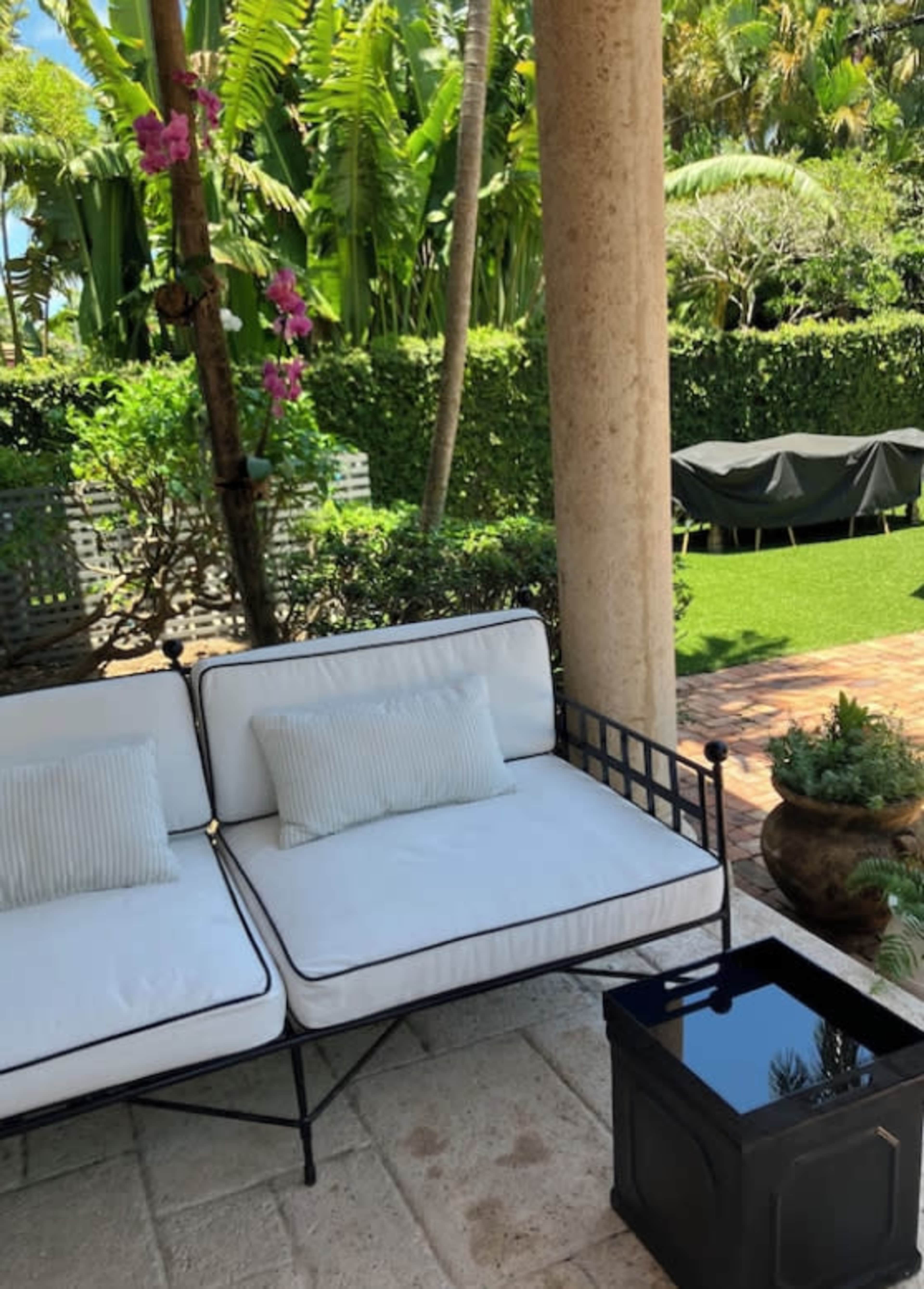A metal-framed white couch with two cushions sits beside a small black table under a covered patio, with greenery and a grill visible in the background.
