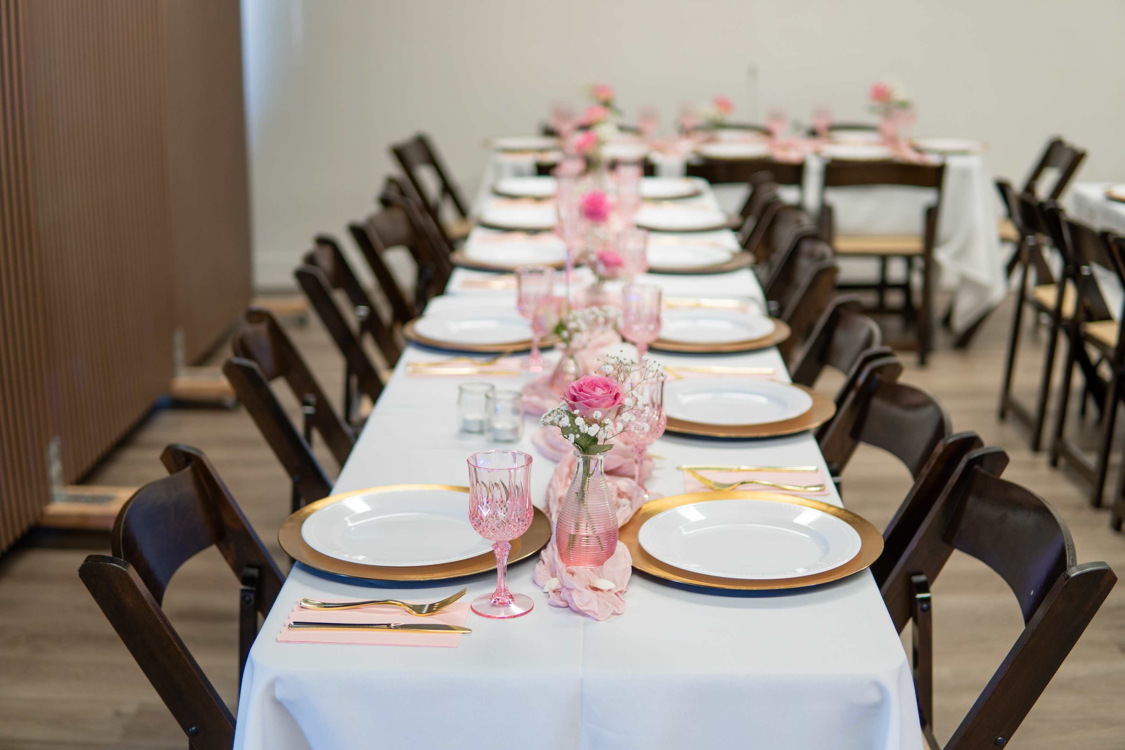 The image shows a banquet setup with multiple tables arranged in a row, each adorned with white tablecloths, gold-rimmed plates, pink glasses, and flower centerpieces.