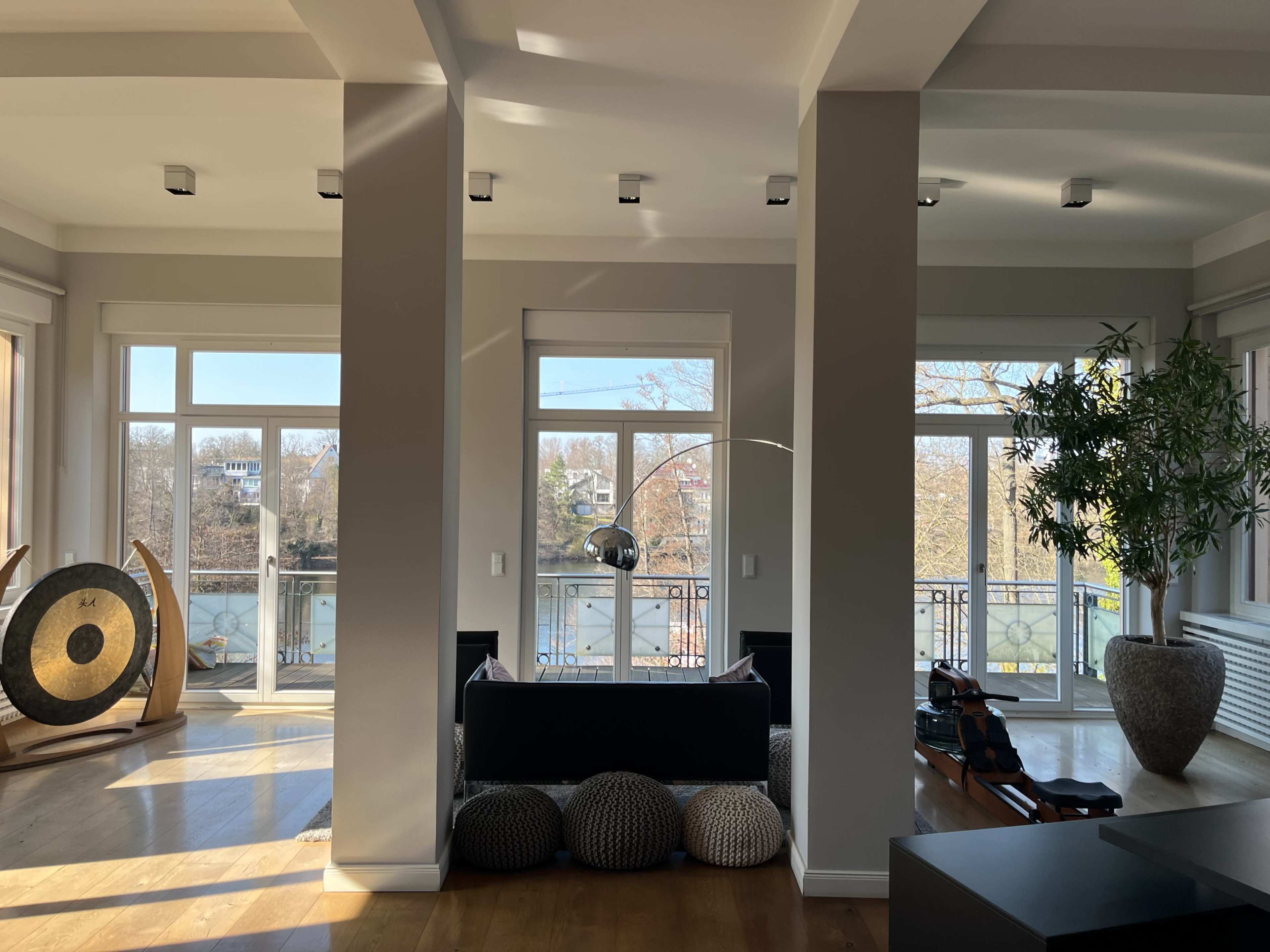 The image shows a modern living room with large windows, hardwood flooring, a black sofa in the center, and decorative items including floor cushions and a plant.