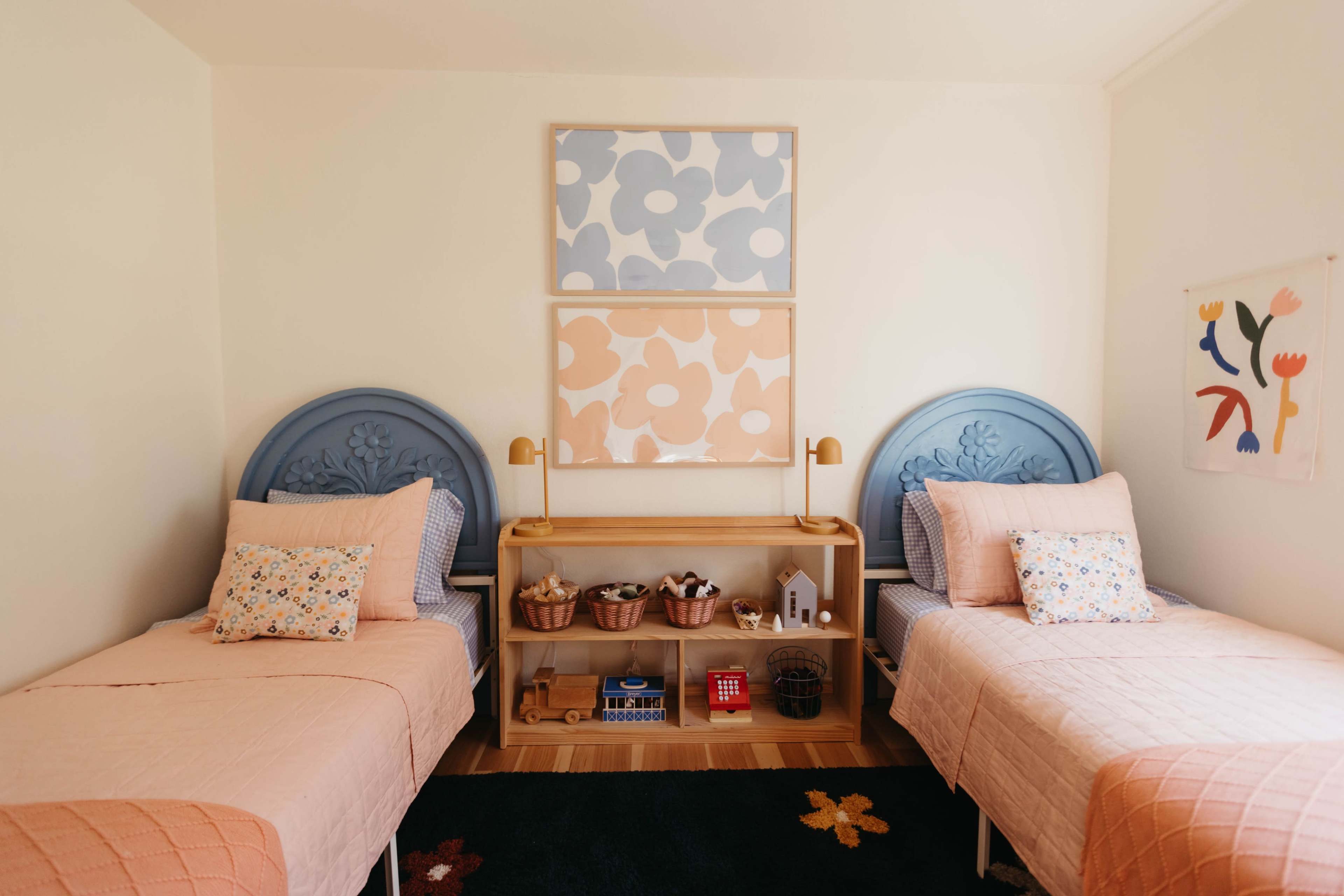 A children's bedroom with two identical beds, each adorned with pastel-colored bedding, and a decorative shelf displaying toys and decor in between.