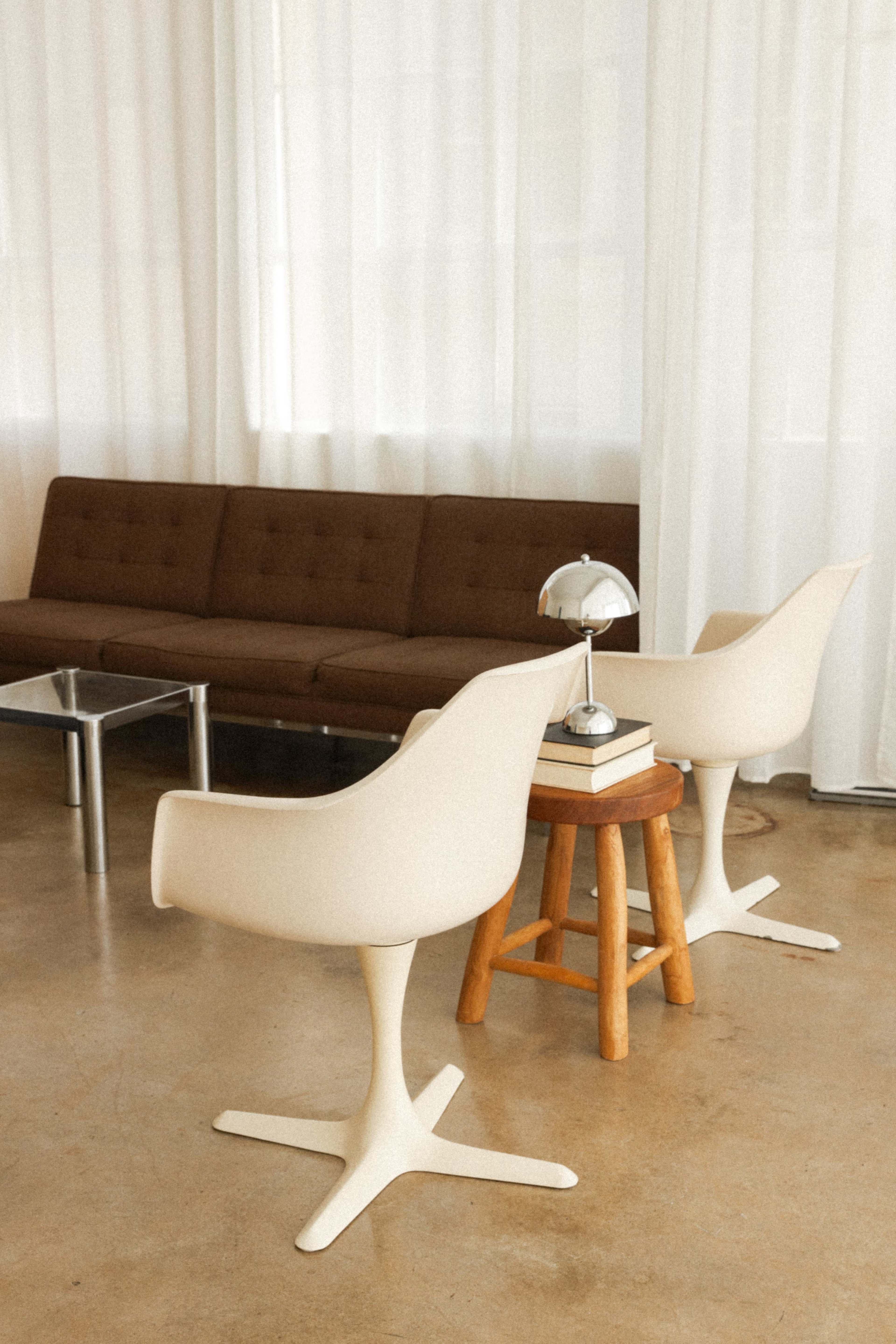A minimalistic living area with a brown sofa, two white chairs, a small wooden stool, and a table with a book and a lamp.