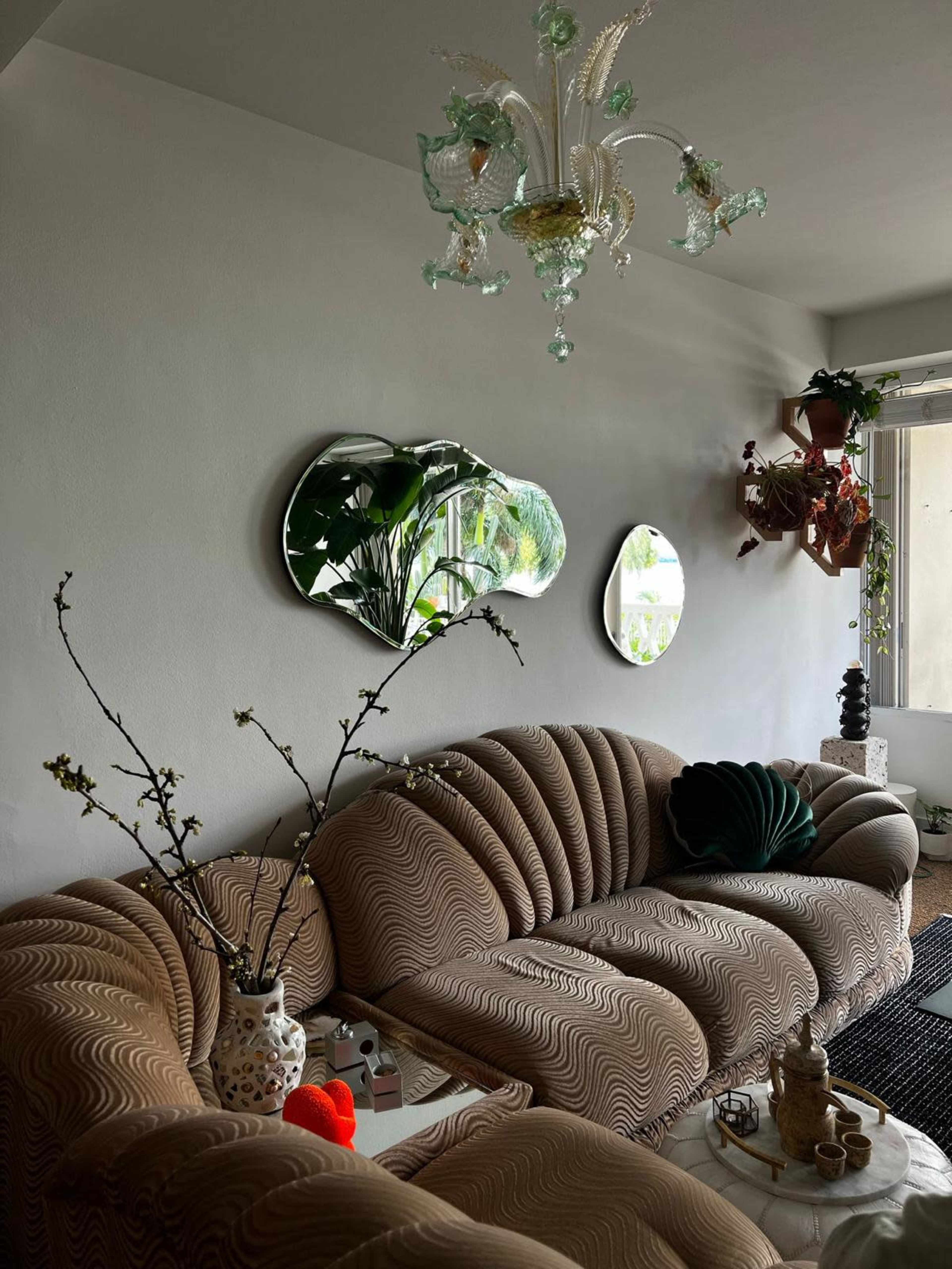 A curved beige sofa is positioned against a wall adorned with two mirrors and plants, complemented by a chandelier and decorative items on a coffee table.