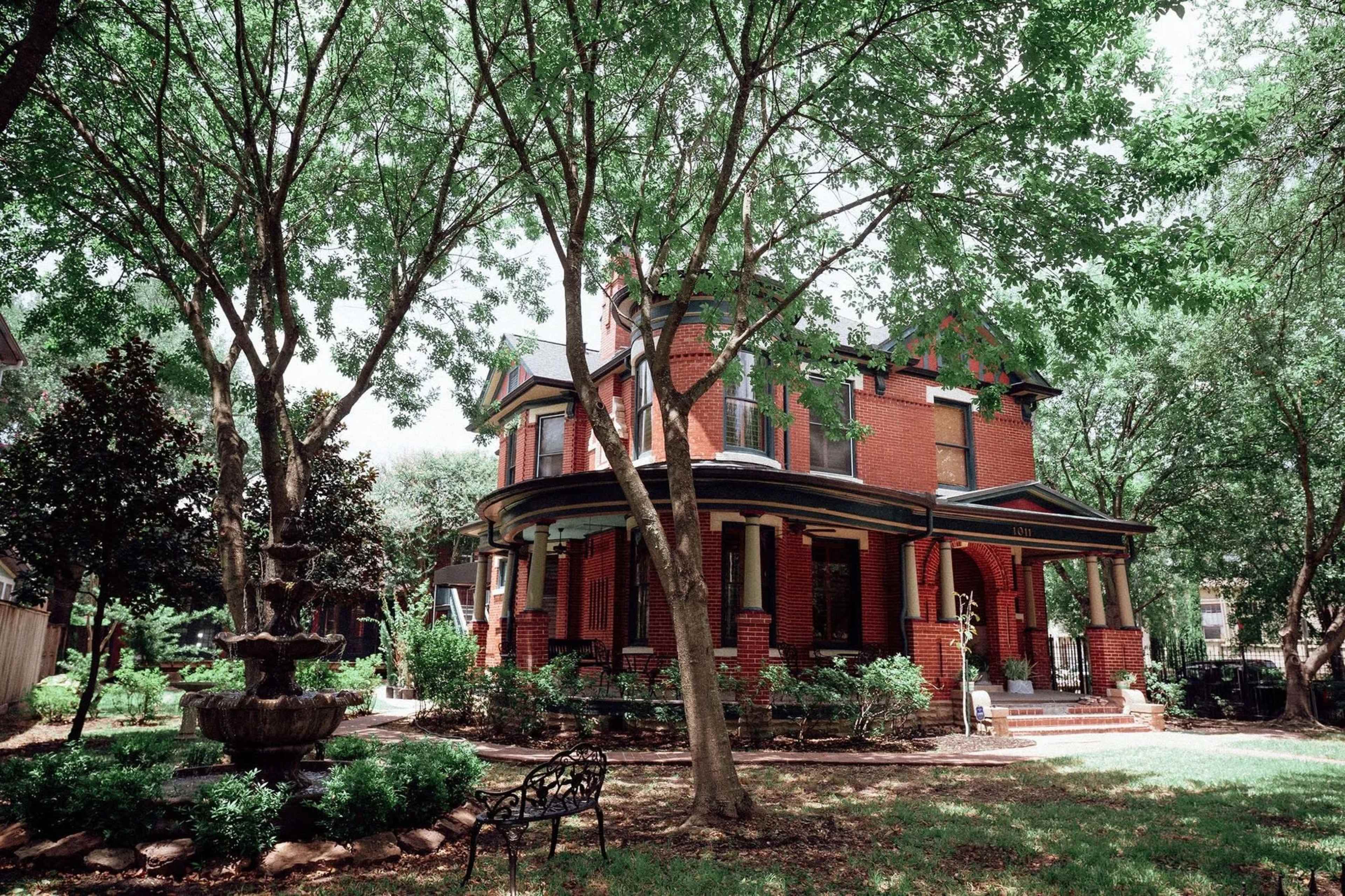 A brick house with a wraparound porch is surrounded by trees and a small garden featuring a fountain.