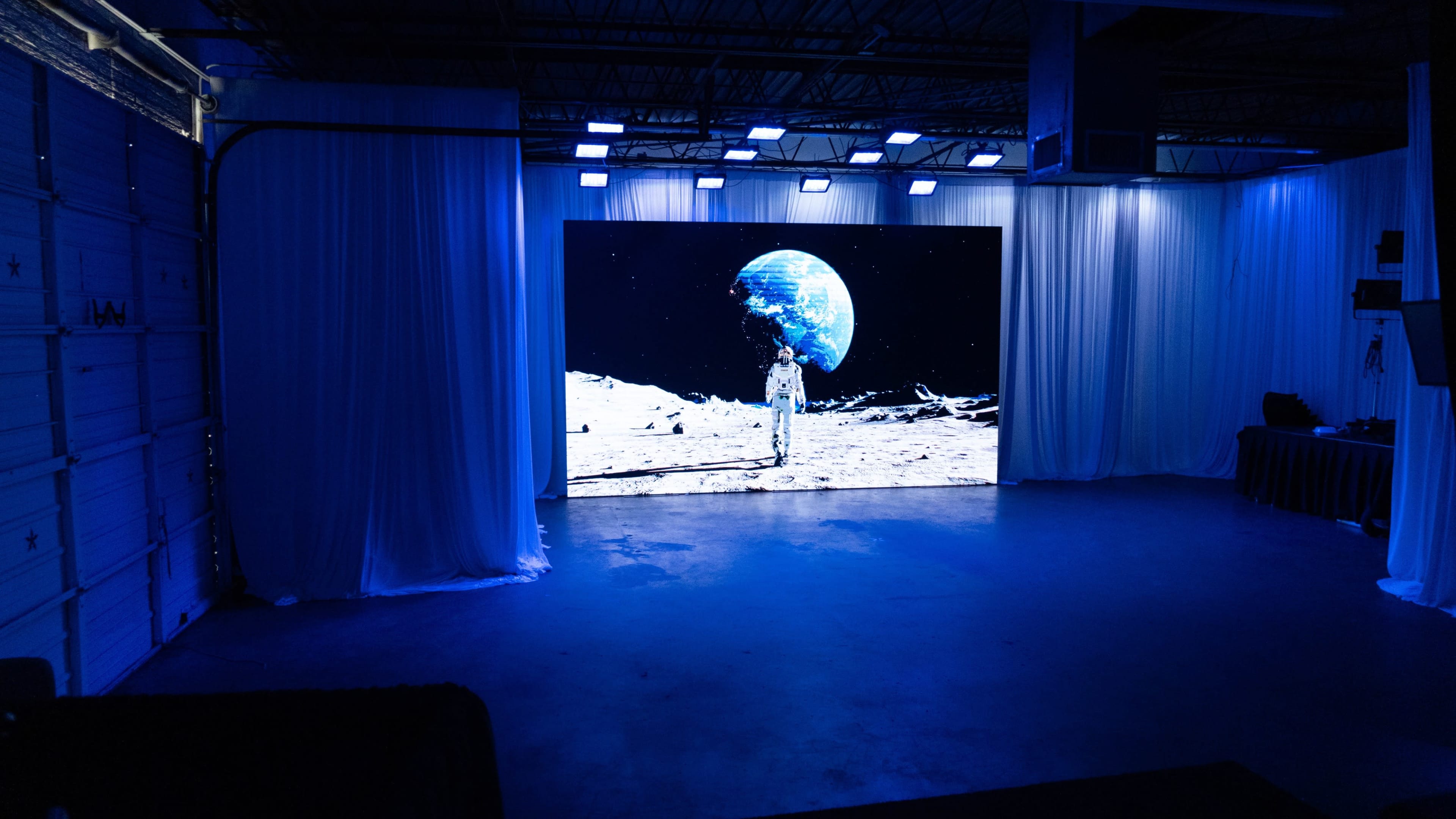 A large screen displays an astronaut standing on the moon with Earth visible in the background, set in a dimly lit, draped room.
