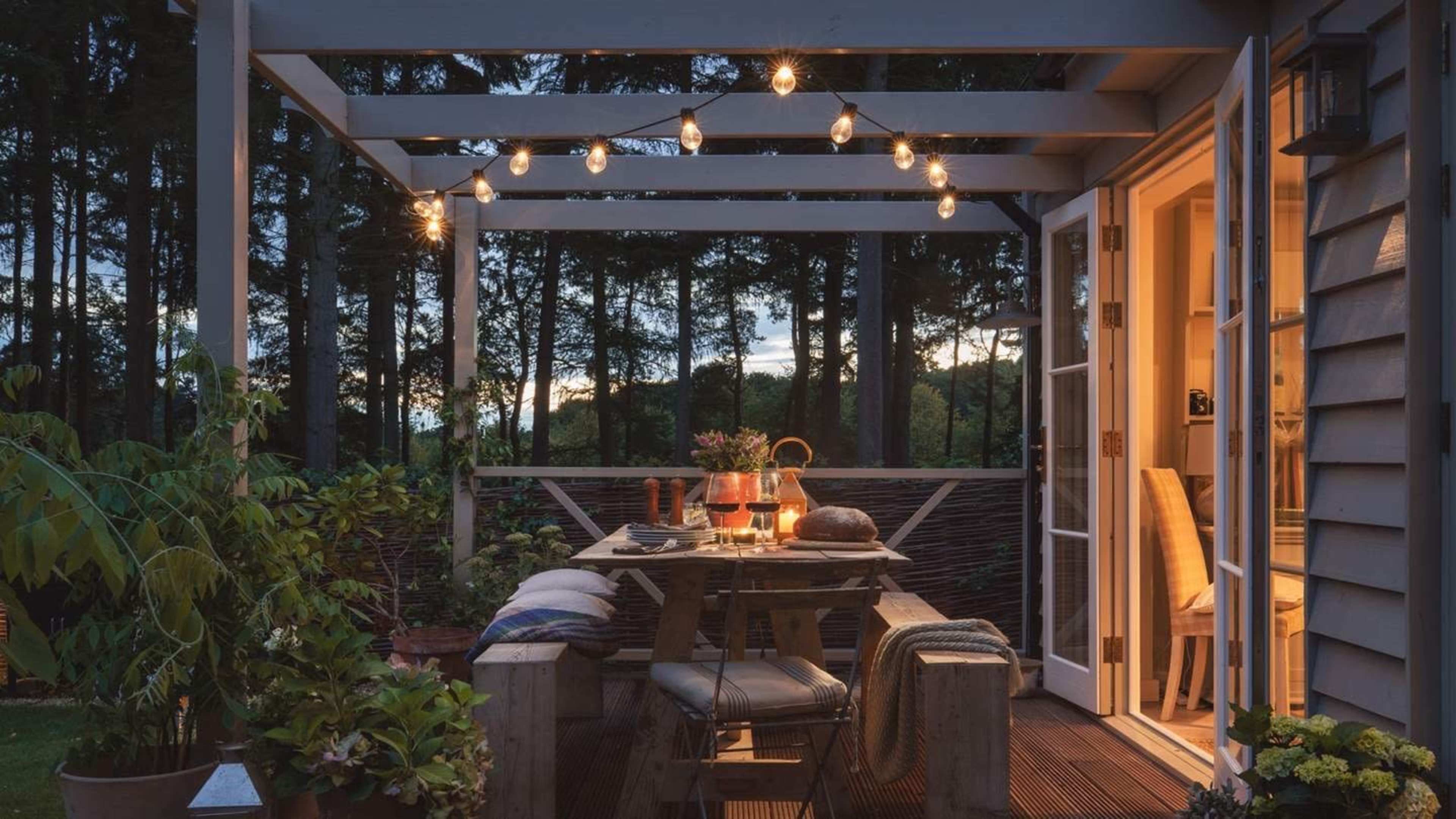A wooden patio with string lights above a dining table set for a meal, surrounded by greenery and trees at dusk.